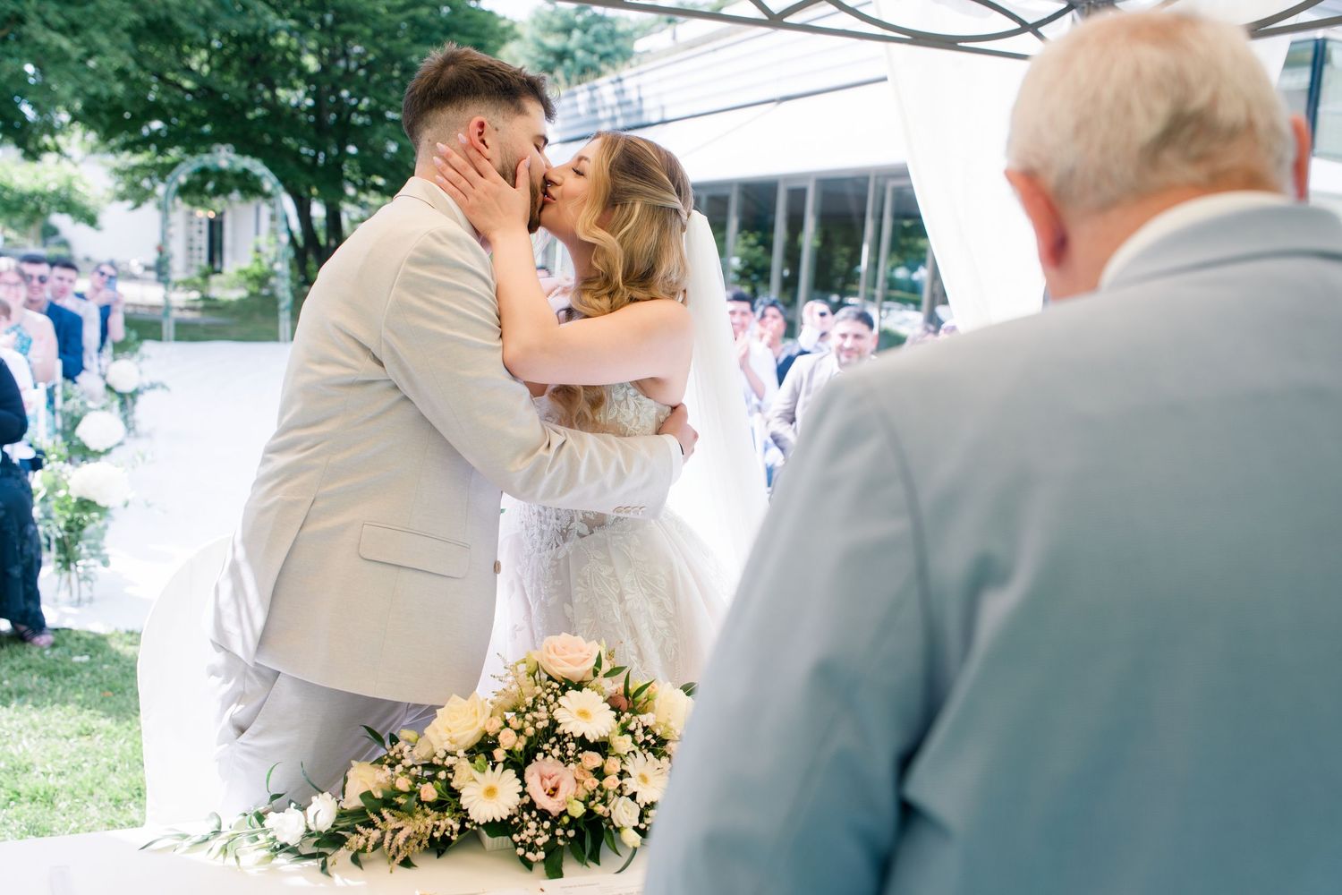 Bride and groom share first kiss at outdoor wedding ceremony with floral bouquet in foreground.