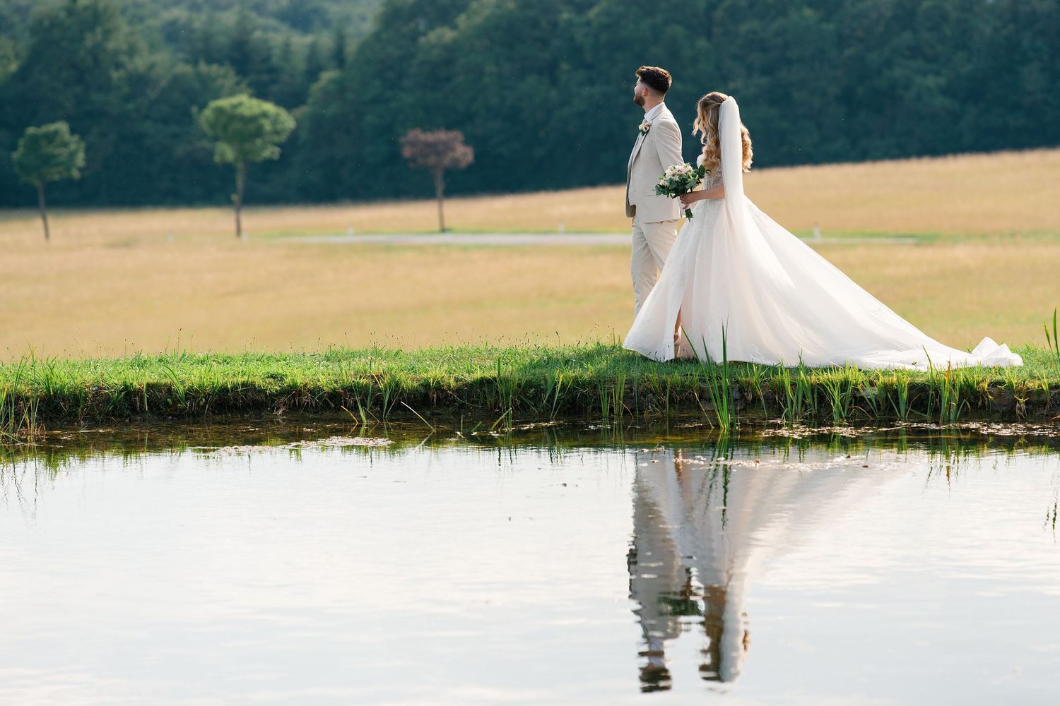 Bride and groom standing by a serene pond with their reflection visible, surrounded by lush green fields.