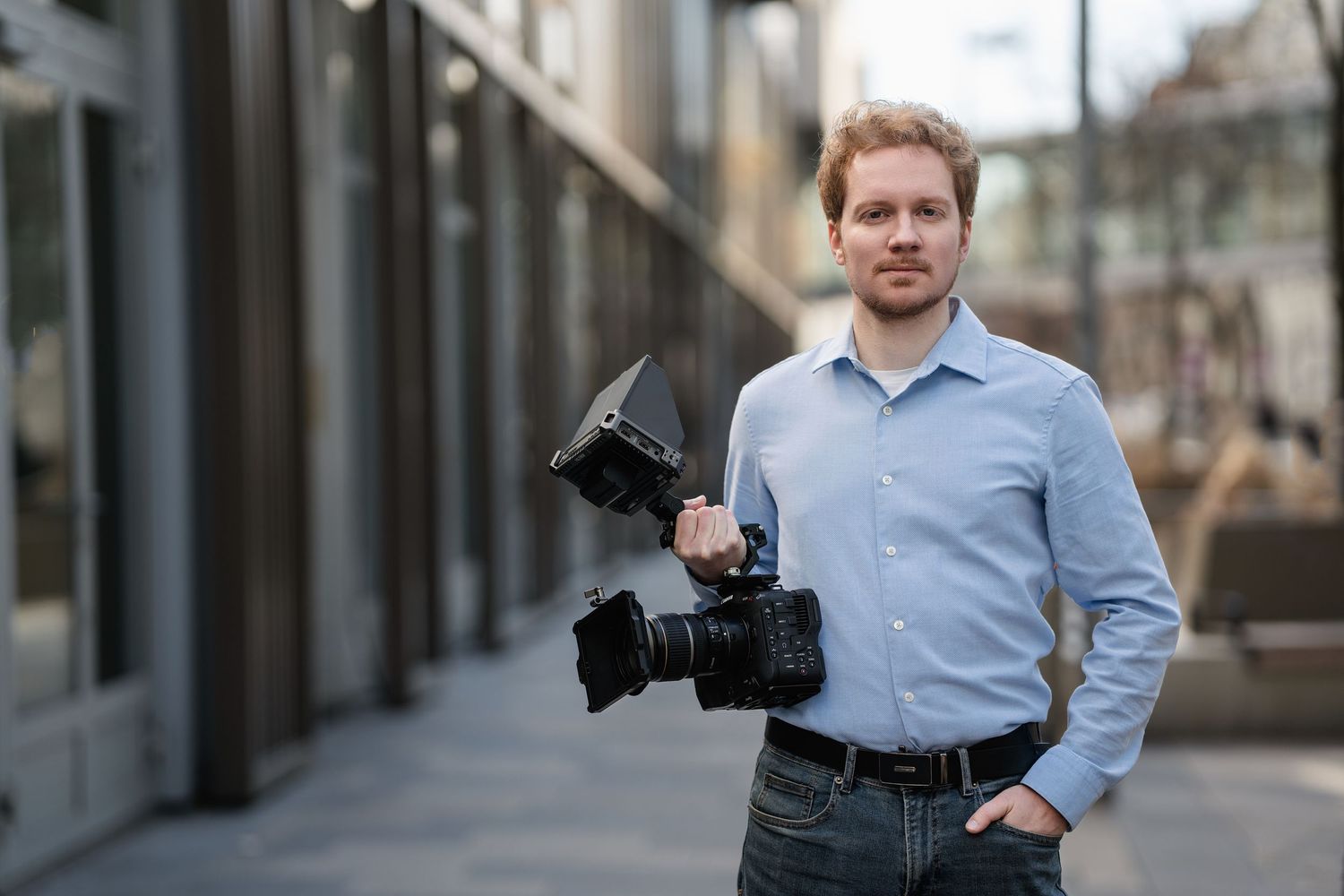 Professional photographer in light blue shirt holding camera gear while standing in urban environment.