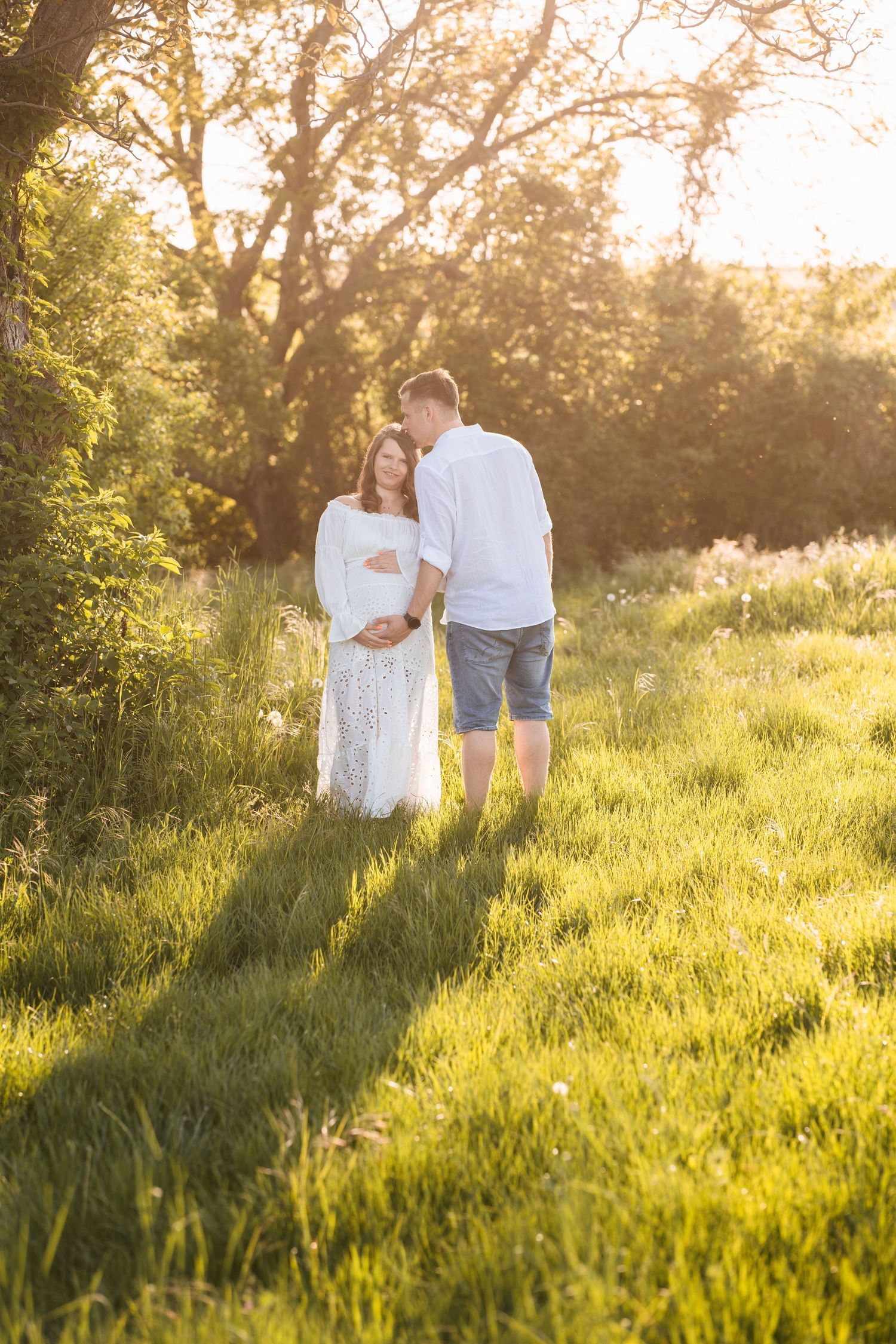 A couple walks hand in hand through a sunlit meadow at golden hour as warm light filters through tall grass.