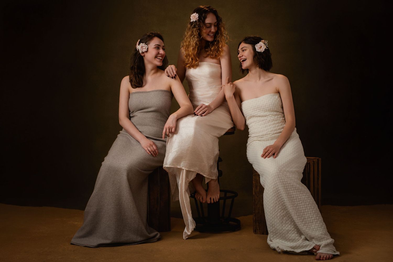 Three people sit together on a dark background wearing elegant dresses in neutral tones.