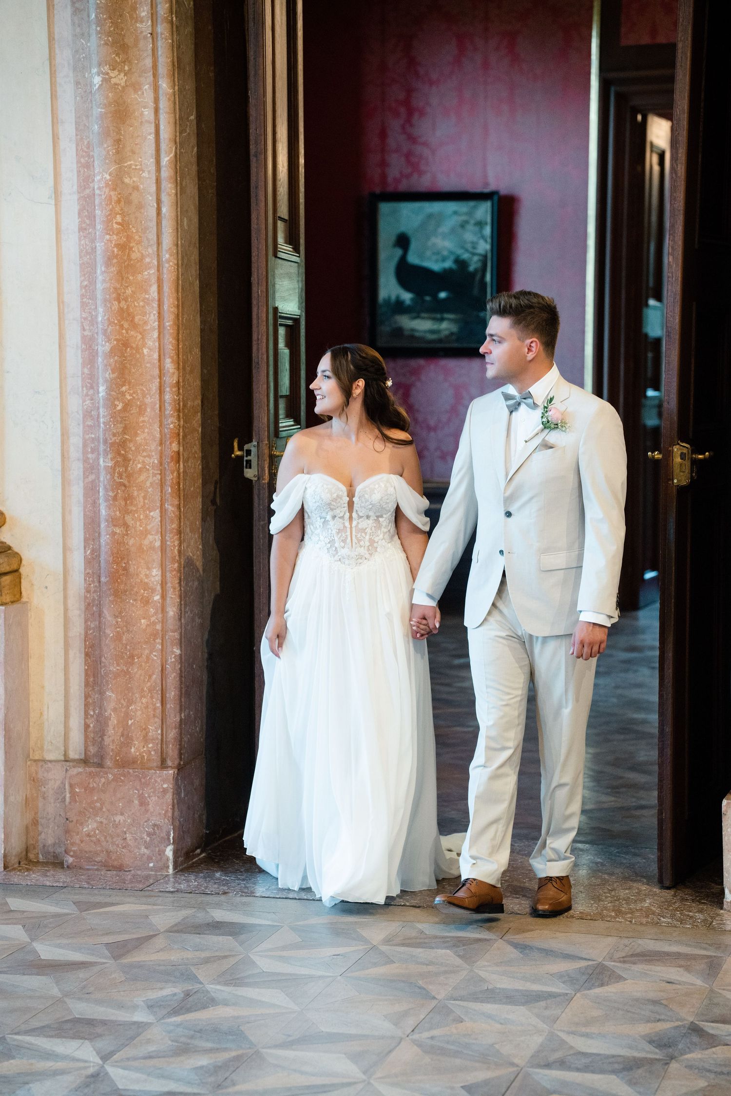 Bride in off-shoulder gown and groom in light suit hold hands in elegant hallway.