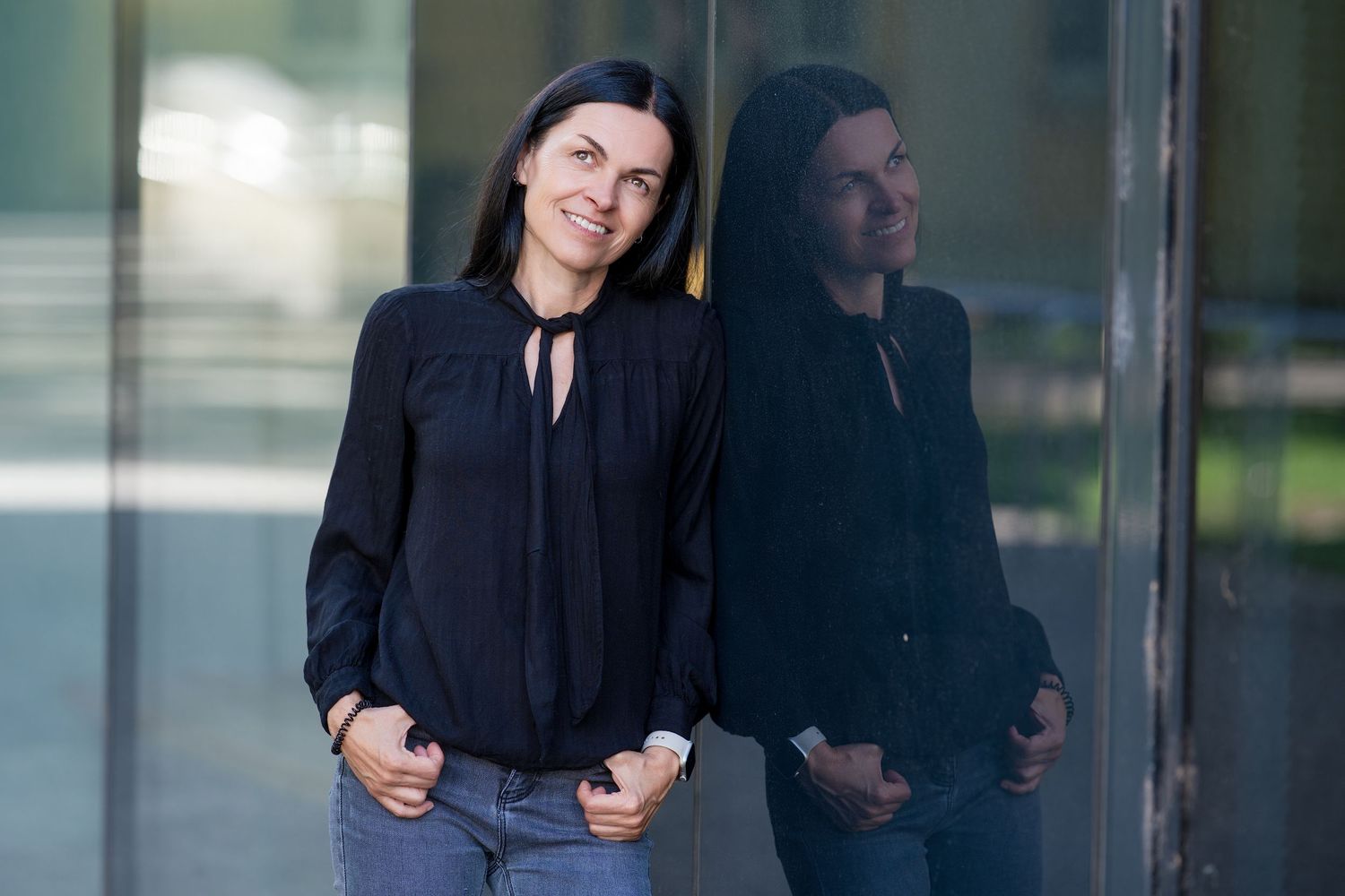 Woman in black blouse leaning against glass wall with reflection, casual professional style outdoor portrait.
