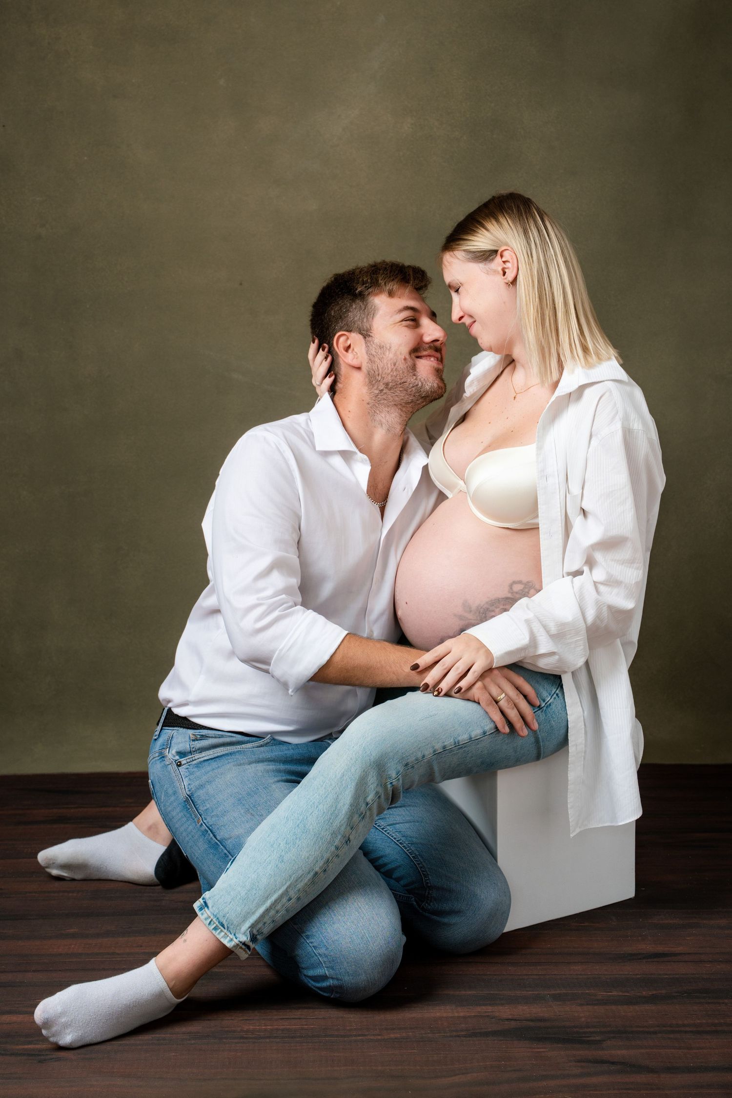 Intimate couple in white shirts and jeans sitting close together on wooden floor in romantic studio portraits.