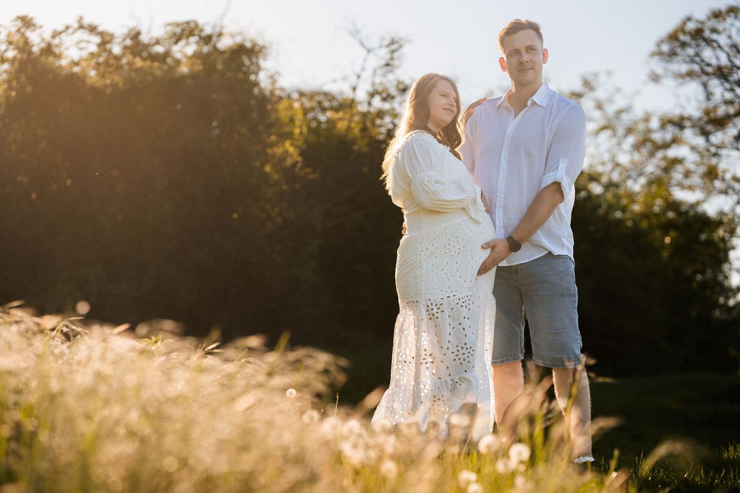 A couple stands together in a sunlit field of tall grass during golden hour at sunset.