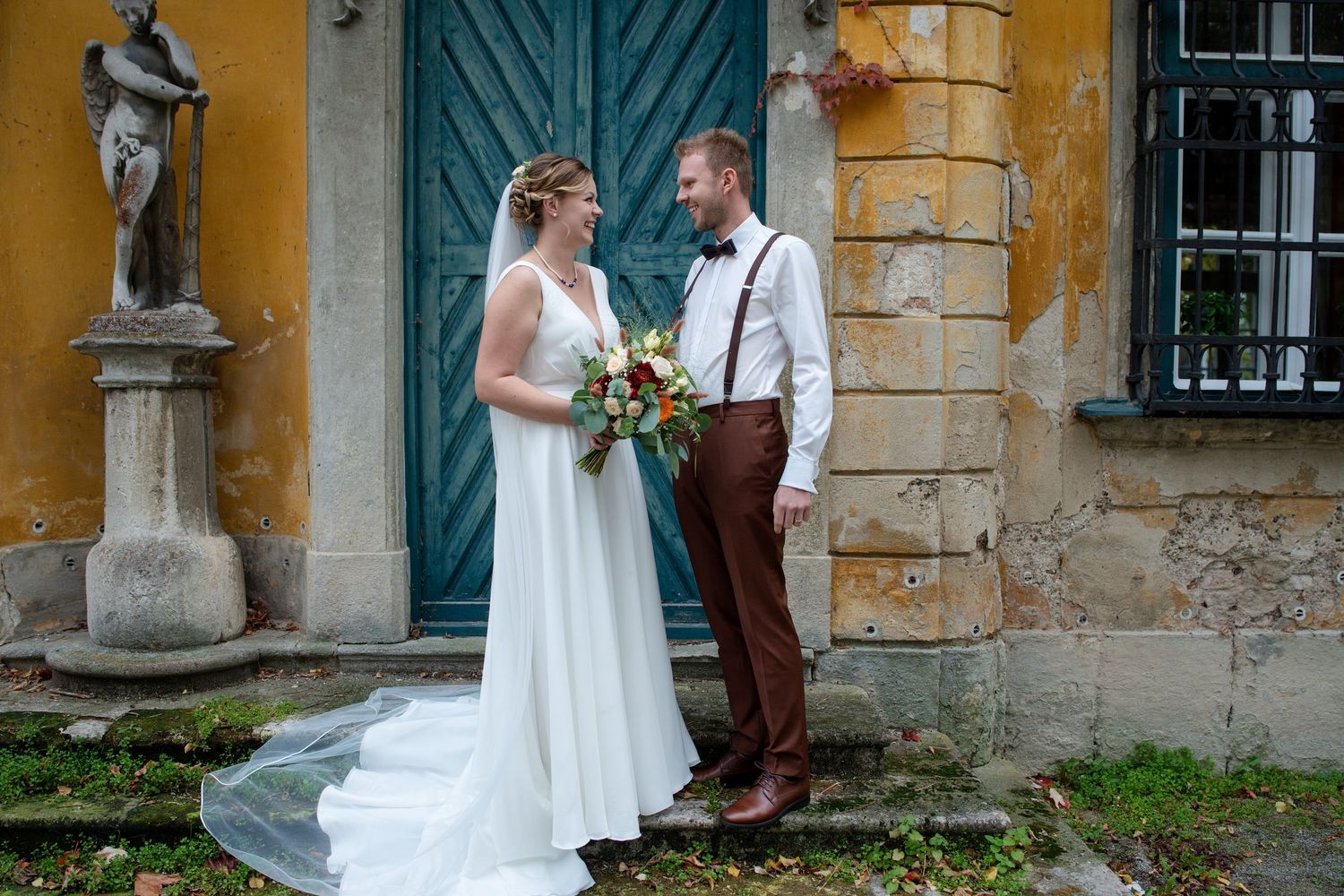 Newlyweds pose for photos against rustic yellow building with teal doors.