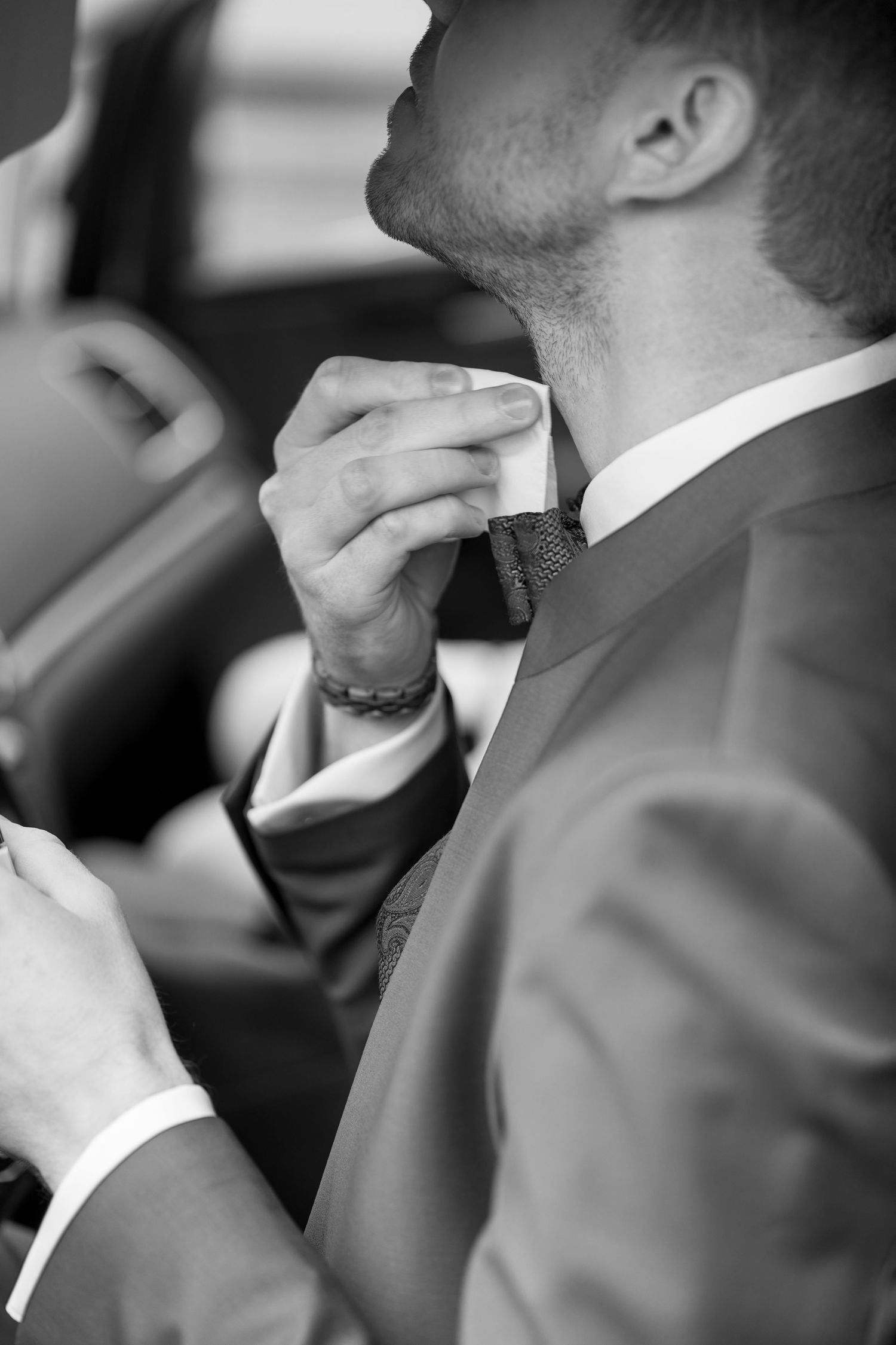 Black and white photo of person in formal attire adjusting their tie in a car.
