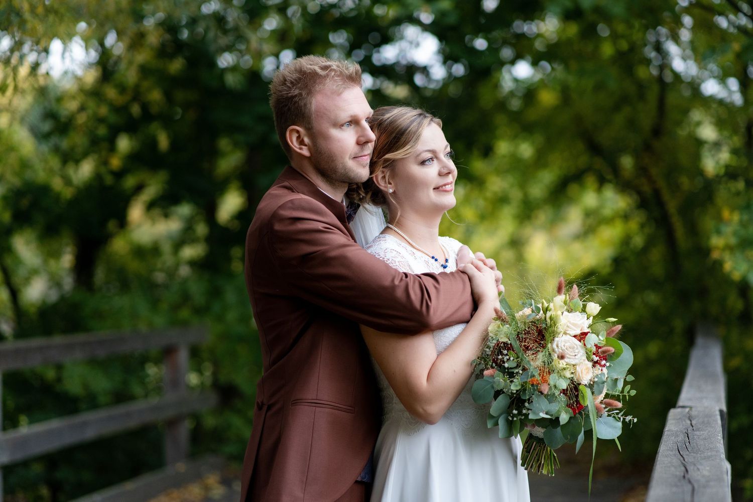 A groom in a brown suit embraces his bride holding a bouquet on a wooden bridge surrounded by lush greenery.