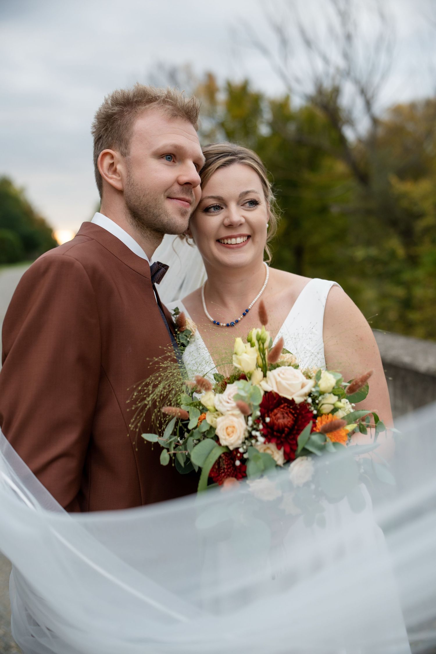 A romantic autumn wedding photo with a flowing veil and burgundy floral bouquet.