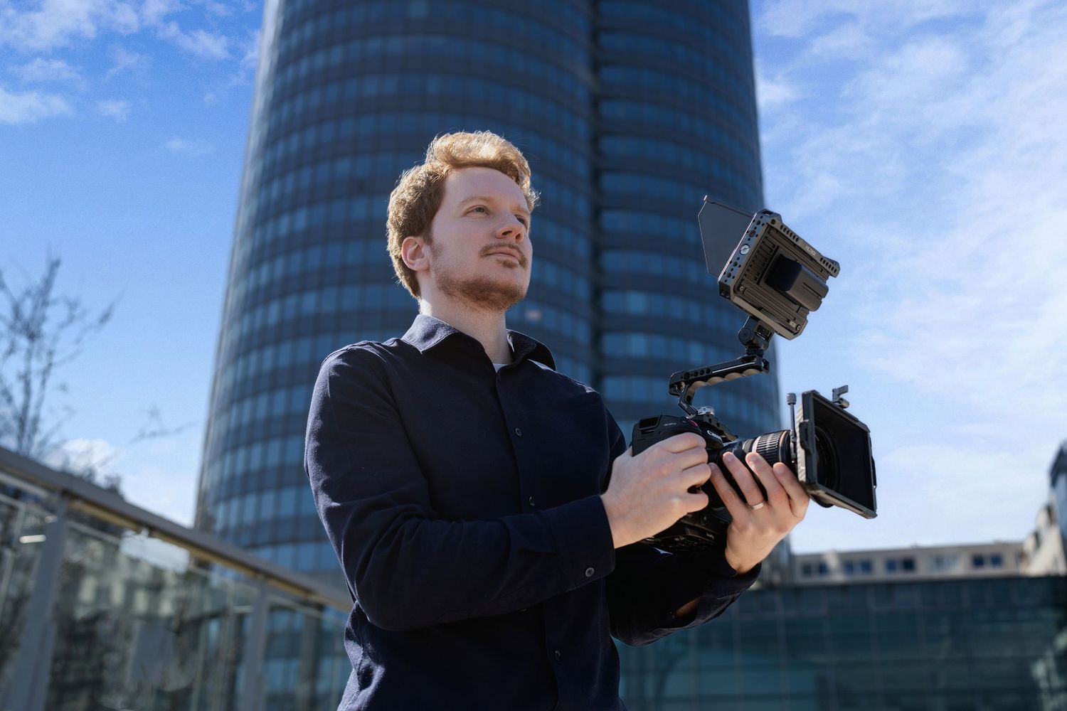 Professional photographer holding camera equipment stands in front of modern glass office building on sunny day.