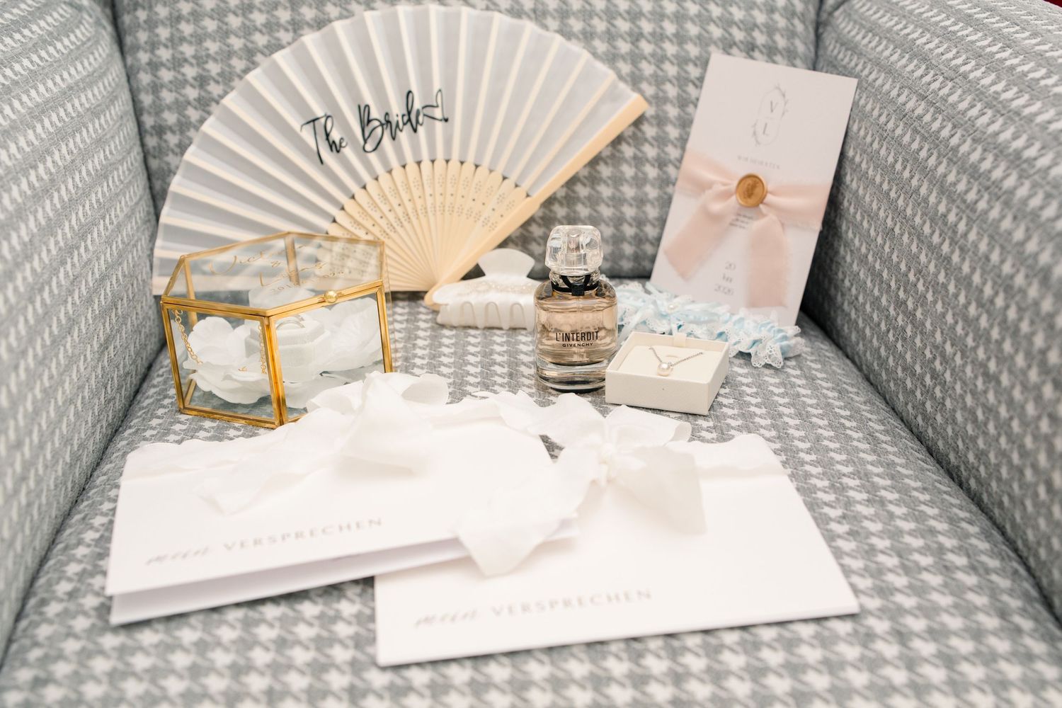 Elegant bridal gift set with fan, perfume, jewelry box, and white stationery on a gray houndstooth chair.