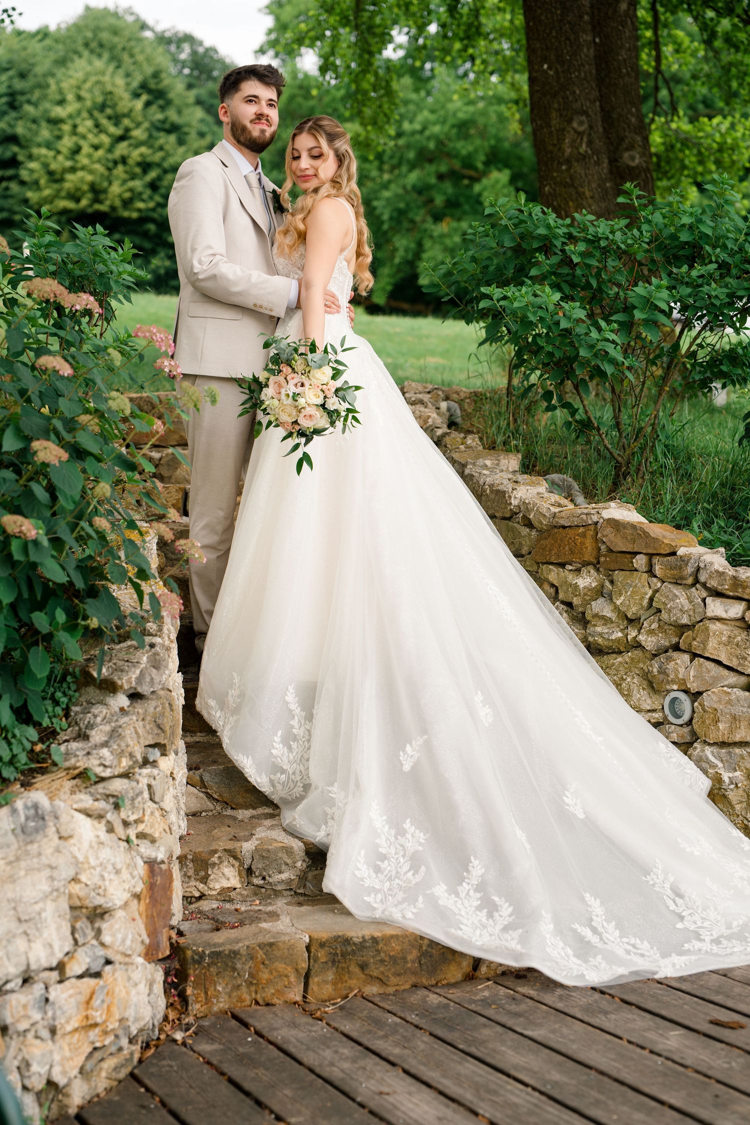 Bride in white lace gown holding bouquet stands with groom in tan suit on stone steps in lush garden.