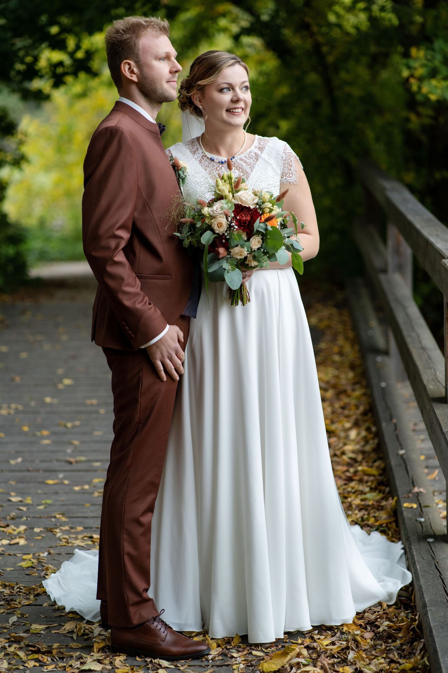 A couple poses on an autumn bridge surrounded by fallen leaves in wedding attire.