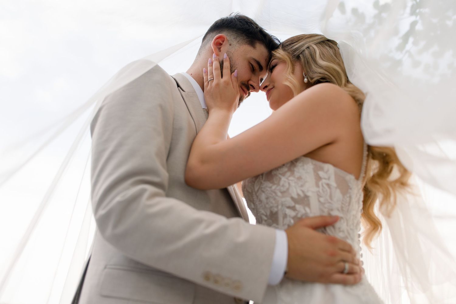 A bride and groom share an intimate moment under a white veil, touching foreheads tenderly on their wedding day.