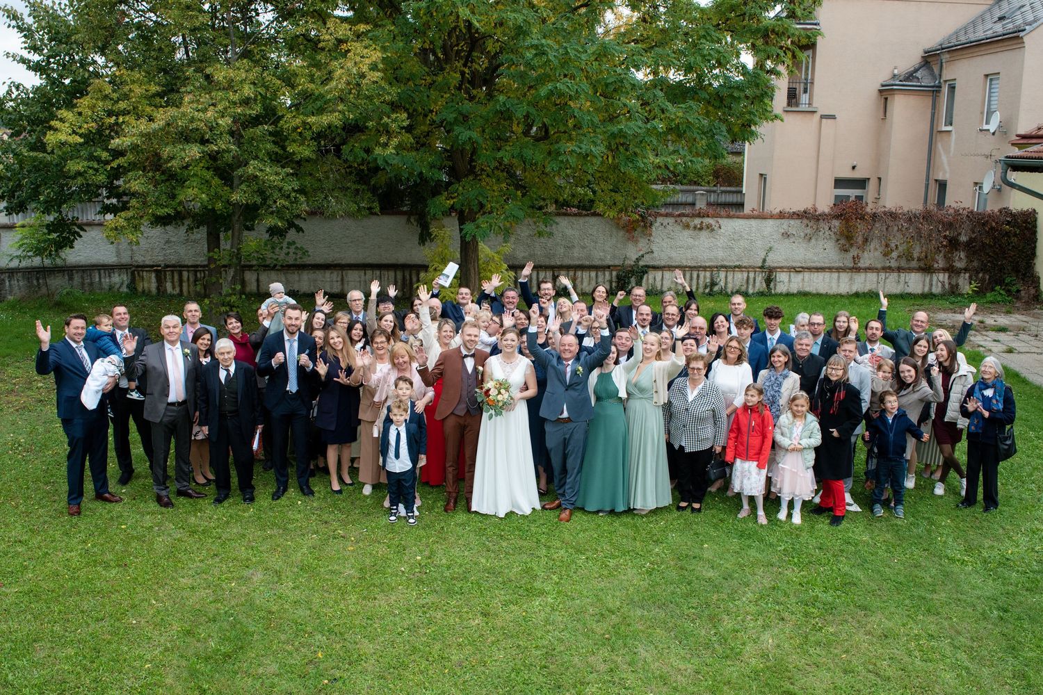 Large wedding party group photo on grass lawn with guests in formal attire.