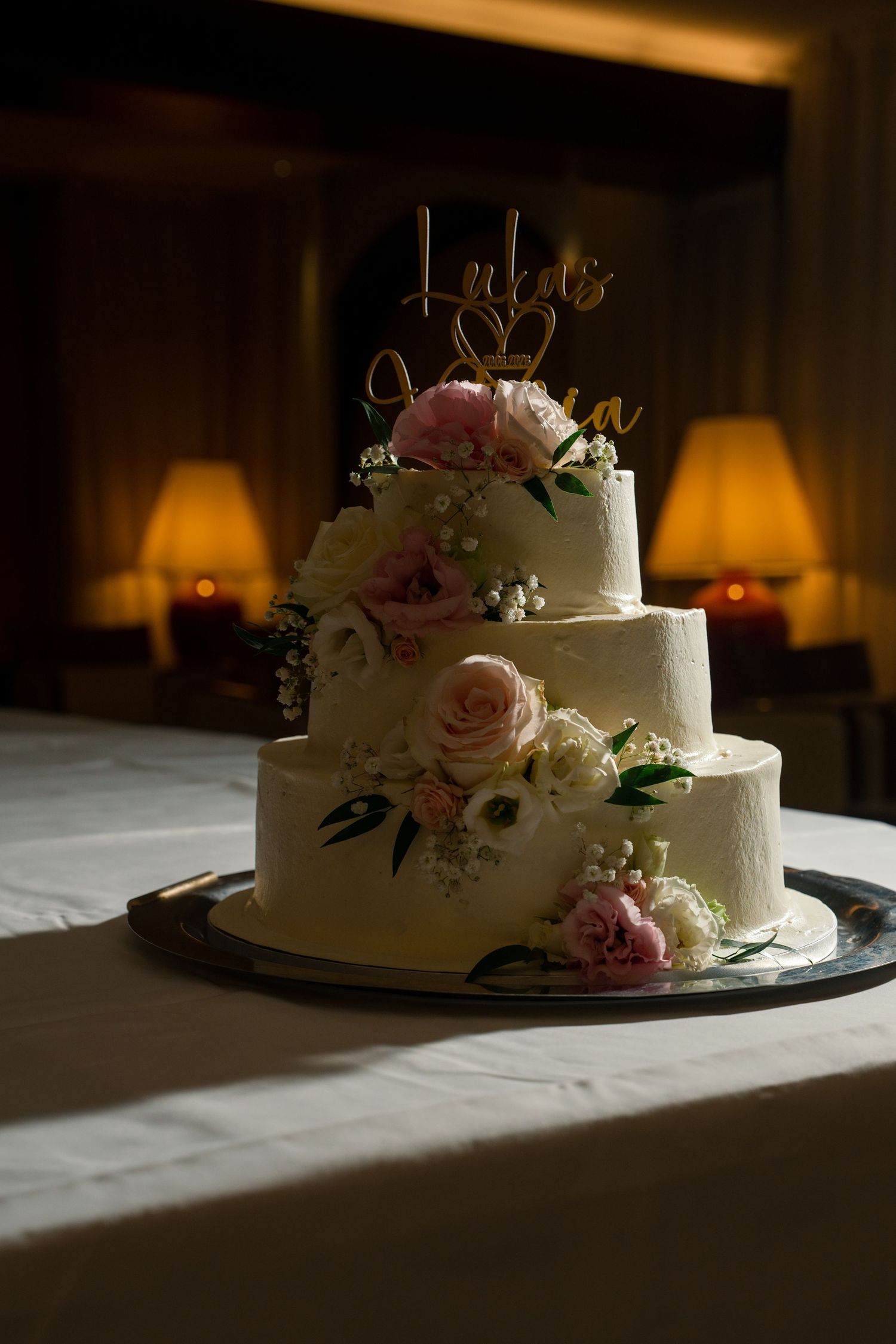Three-tier white wedding cake with pink roses and gold name topper displayed on table with warm candlelight background.