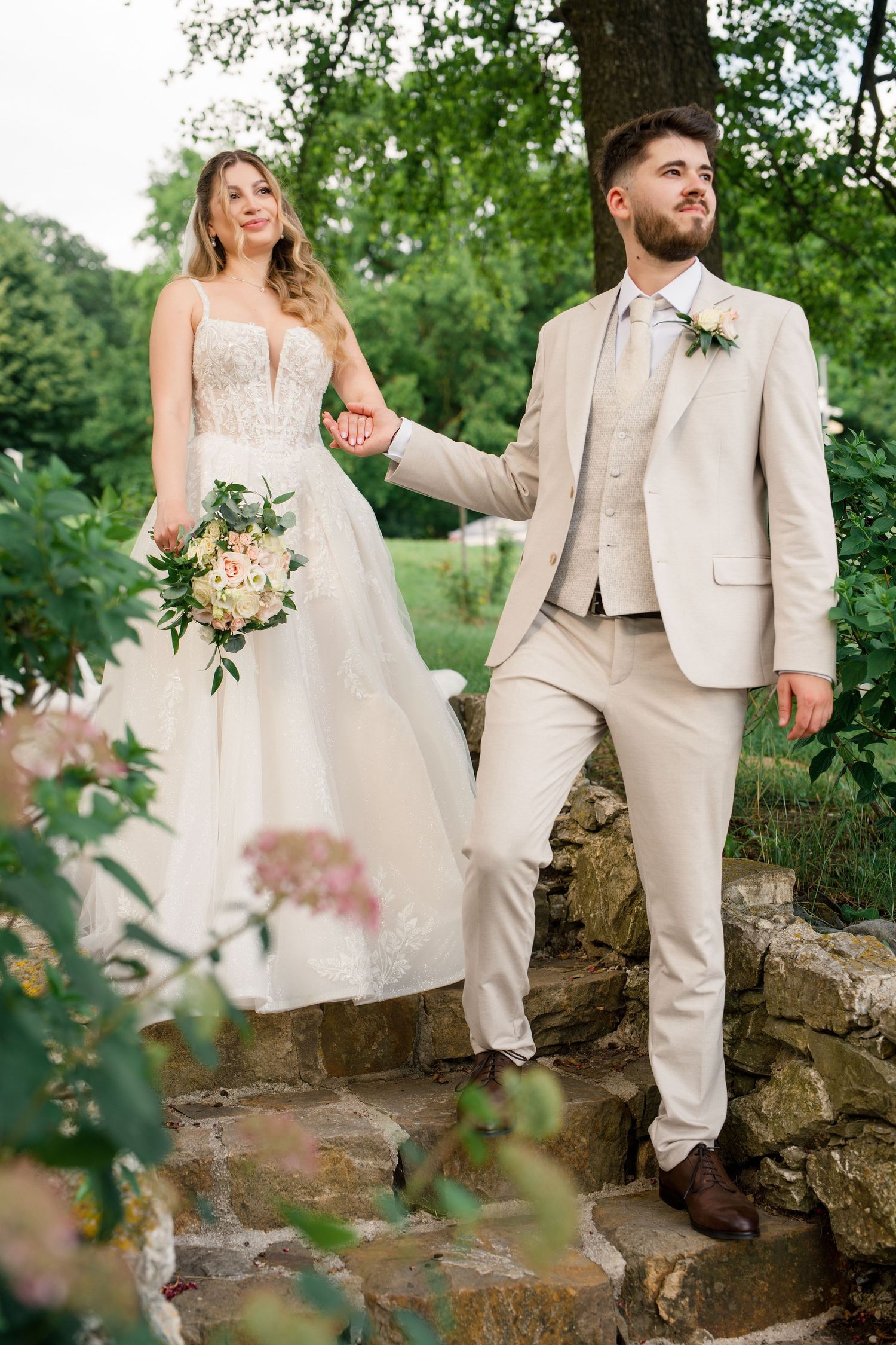 Bride and groom holding hands on stone steps outdoors, she in white lace gown with bouquet, he in beige suit.