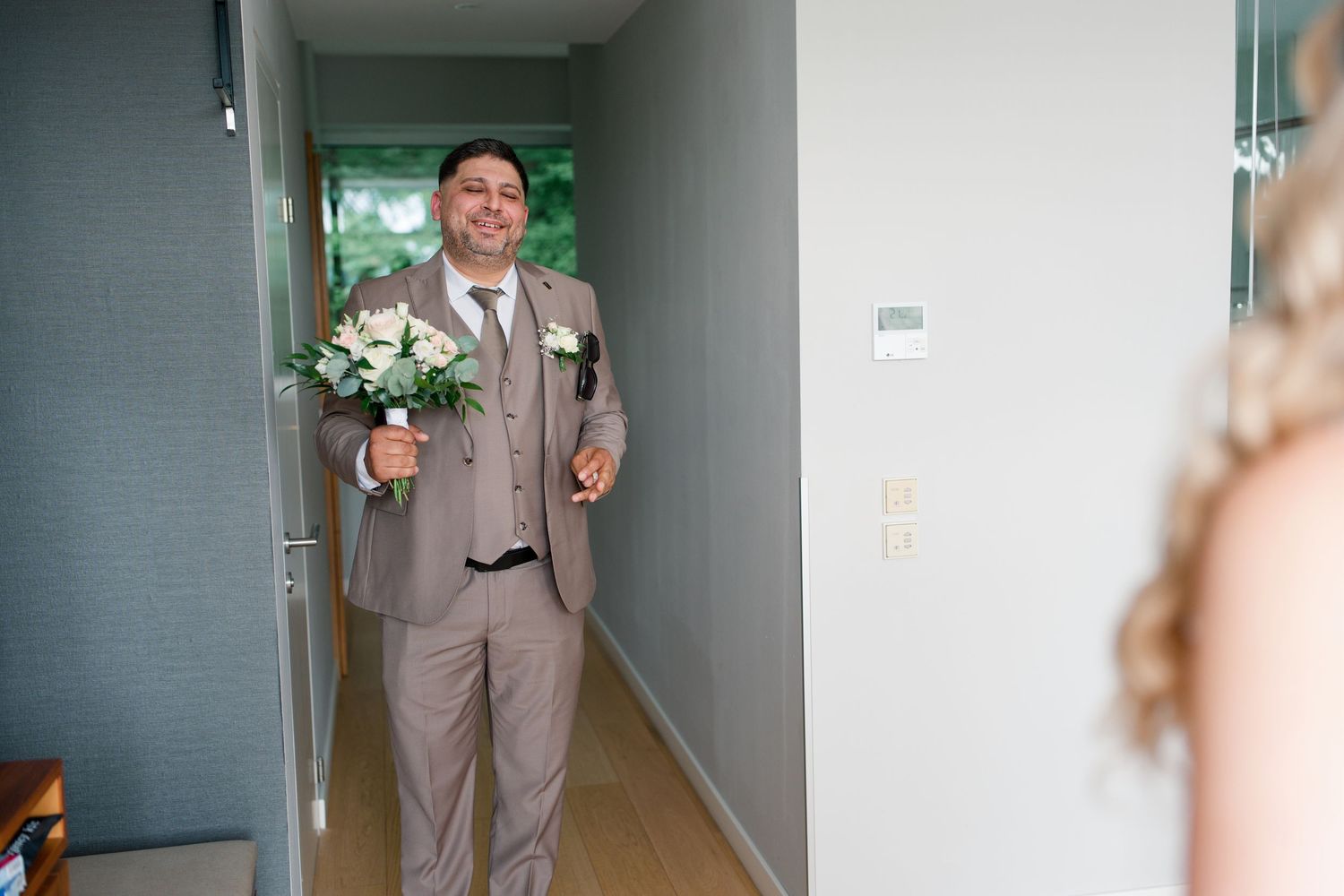 Groom in tan suit holding bridal bouquet smiles at bride during wedding day first look moment.