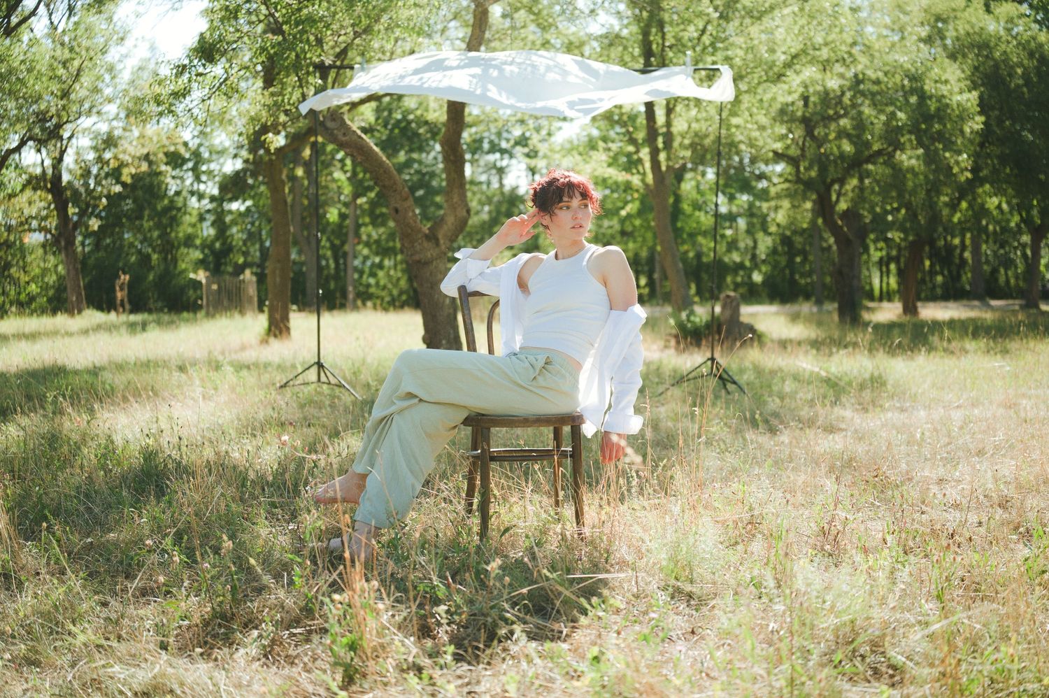 Person in white outfit sitting on chair under umbrella in sunny green meadow surrounded by trees.