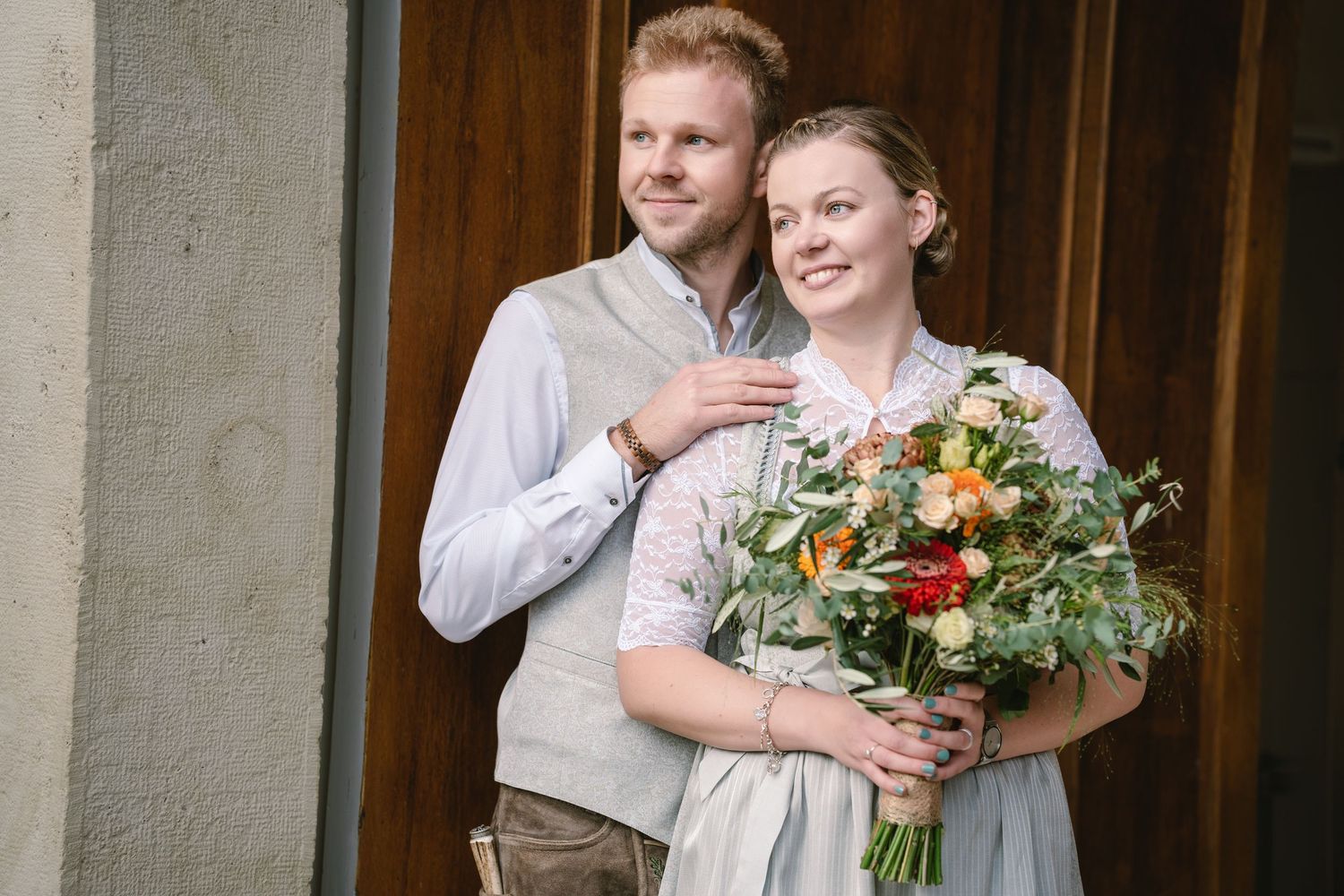Joyful wedding party members laugh together while holding floral bouquets outside a stone building.