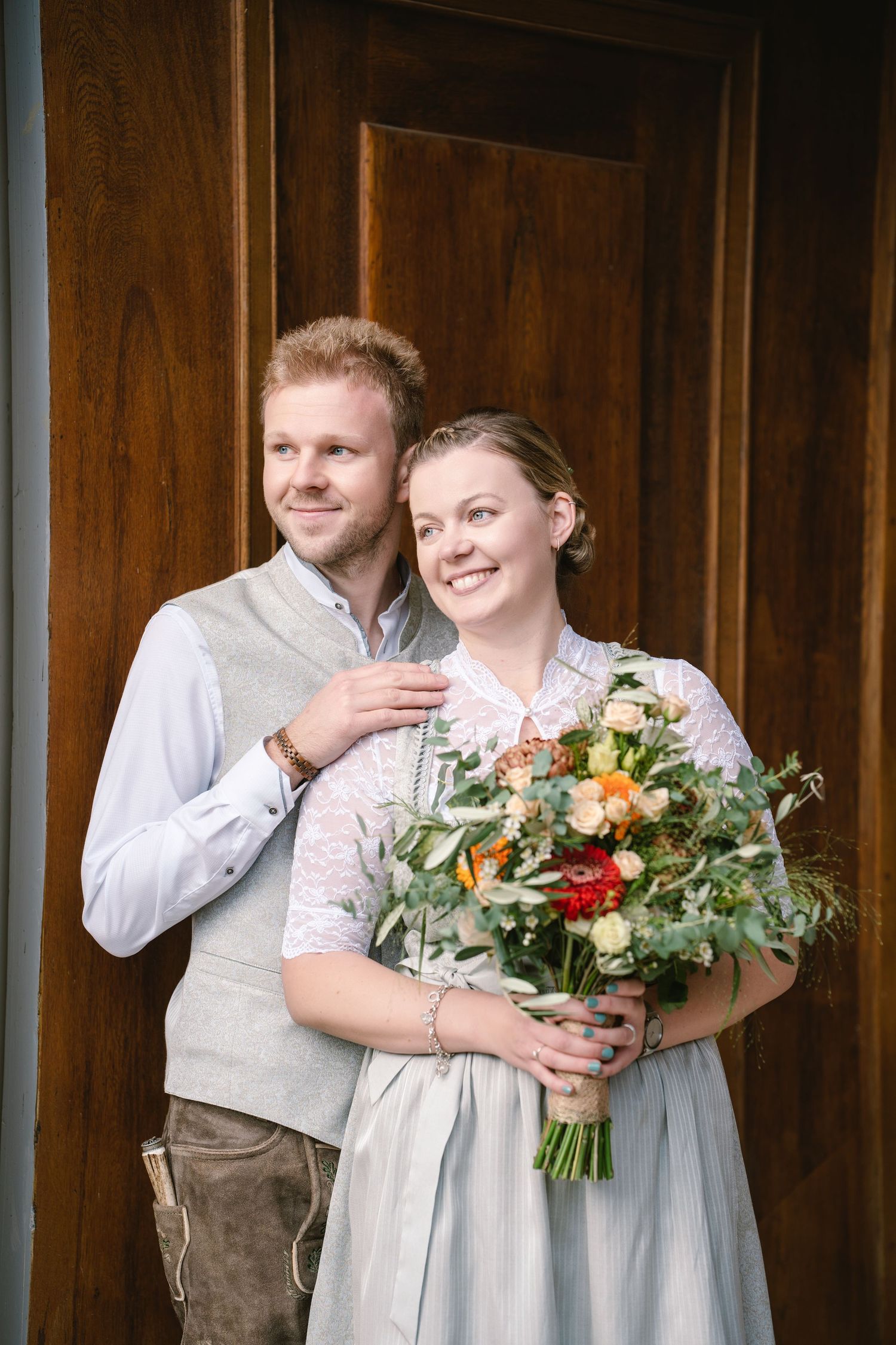 Wedding party members in traditional Bavarian attire hold colorful floral bouquets against a wooden wall backdrop.