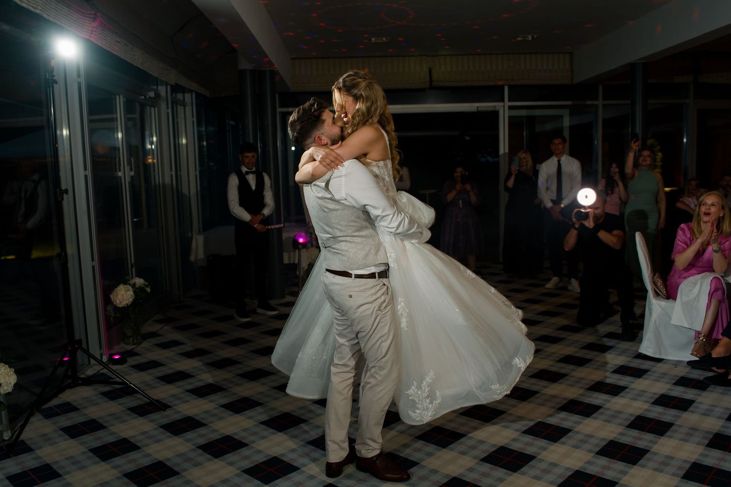 Groom lifting bride during first dance at wedding reception on black and white checkered dance floor.