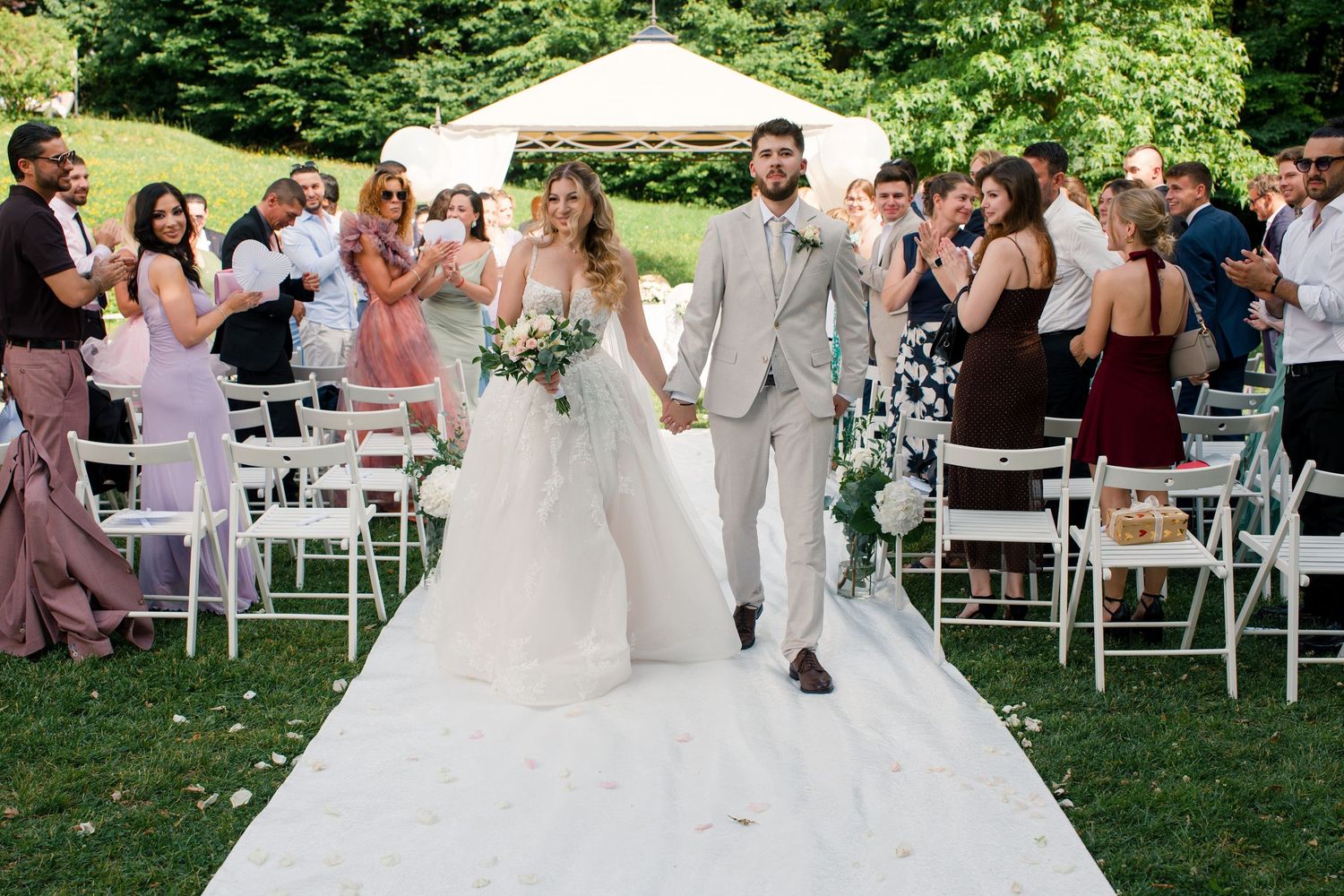 Bride and groom walk down outdoor aisle after wedding ceremony, surrounded by cheering guests on a sunny day.