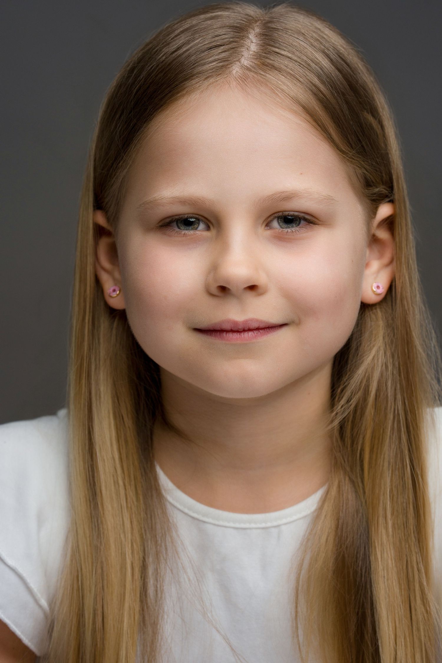 Portrait of a young child with long blonde hair wearing a white t-shirt against a dark gray background.