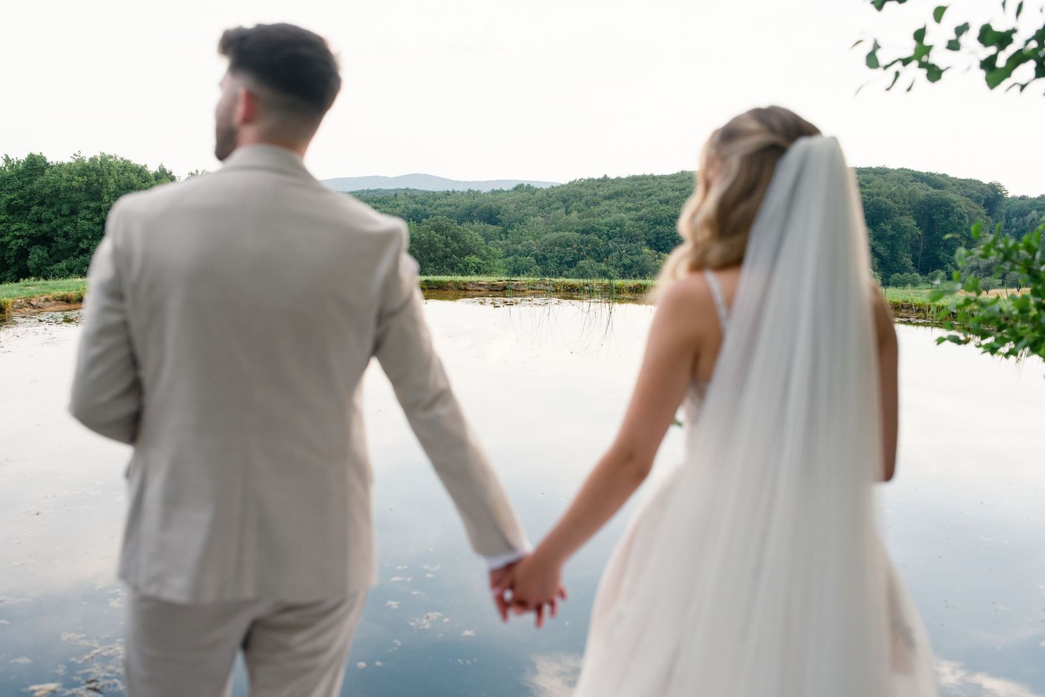 Bride and groom holding hands overlooking a serene lake and lush green hills on their wedding day.
