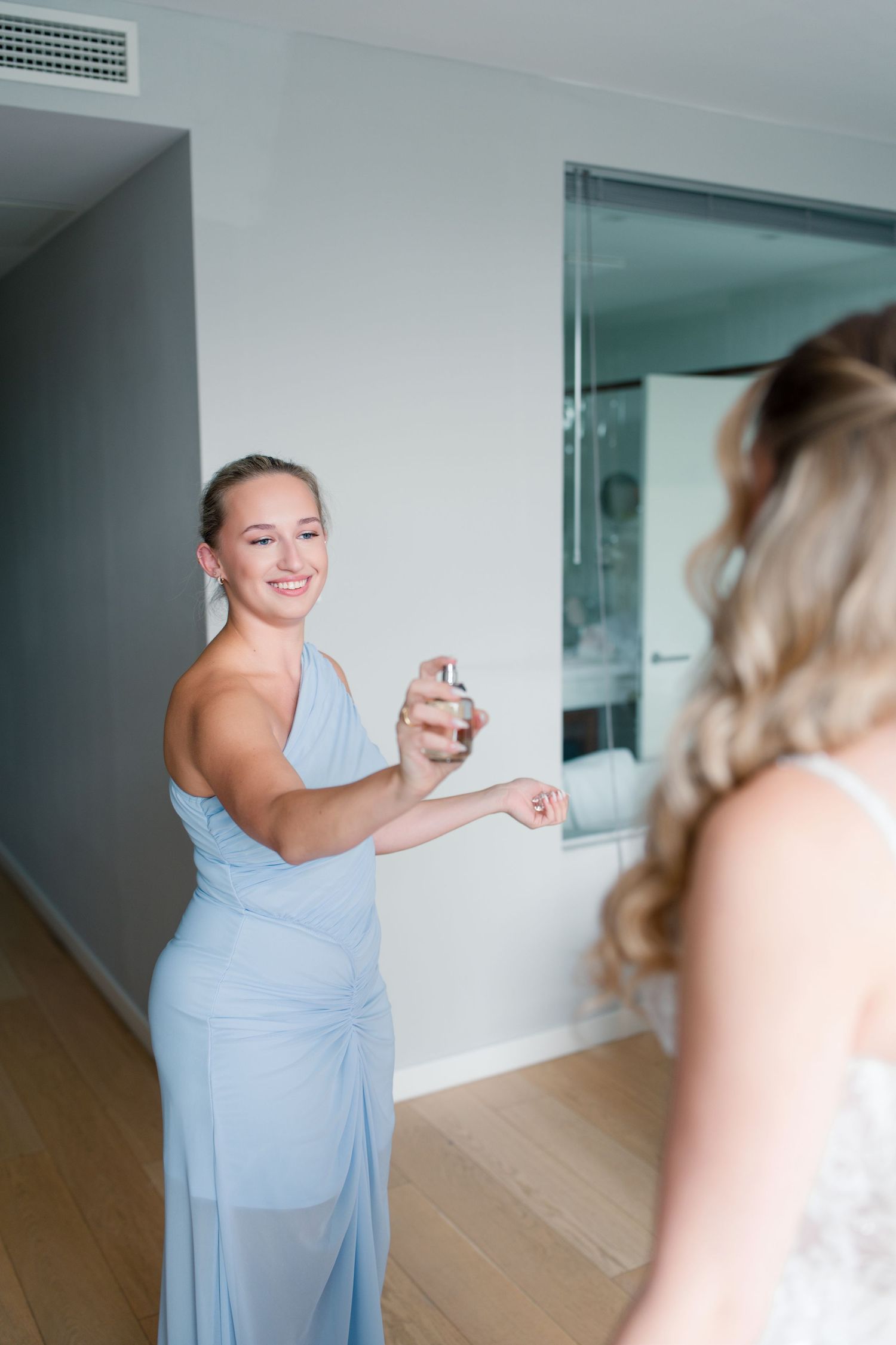 Bridesmaid in light blue dress spraying perfume on bride during wedding preparations in bright modern room.