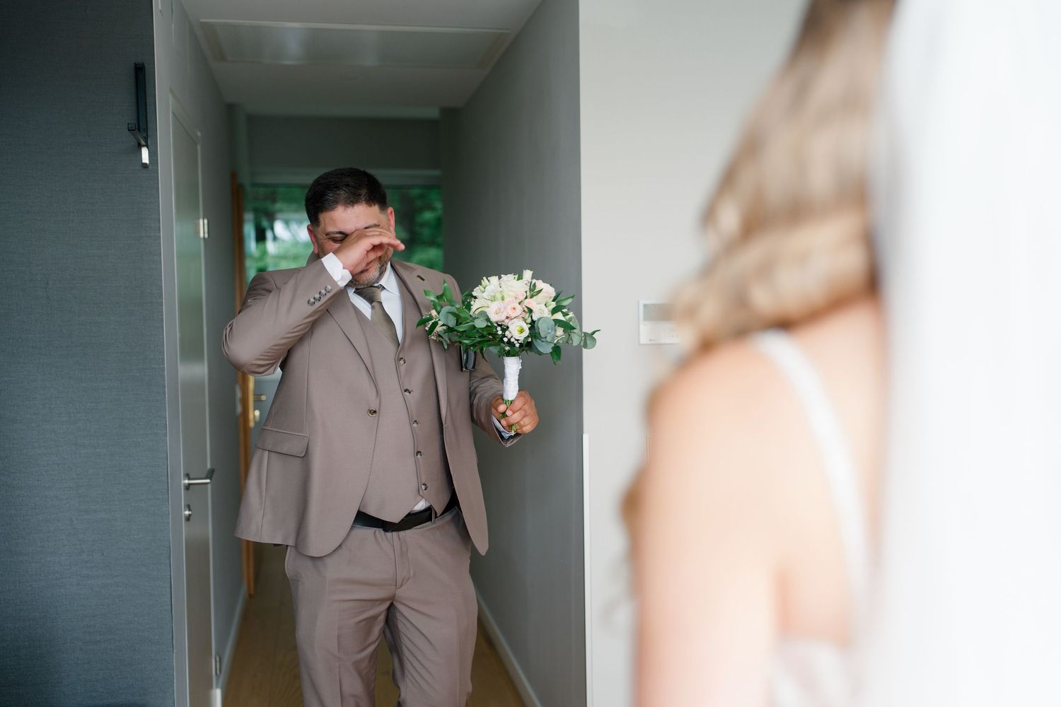 Emotional groom wiping tears holding bouquet as bride appears in doorway during wedding first look moment.