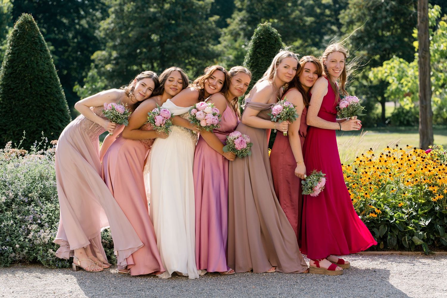 Bridesmaids in pink and red dresses pose together outdoors in a garden setting.