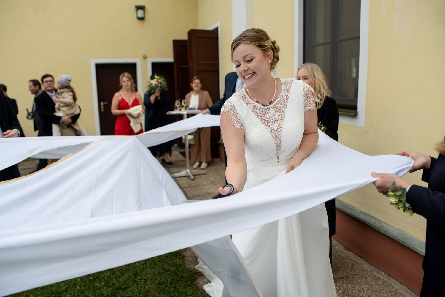 A flowing white wedding veil is held up and spread out by several hands against a beige building exterior.
