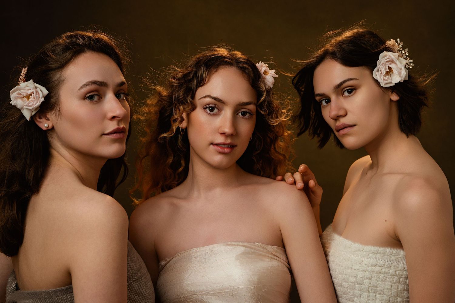 Three women in elegant bridal gowns pose together in a romantic indoor setting with floral accents.