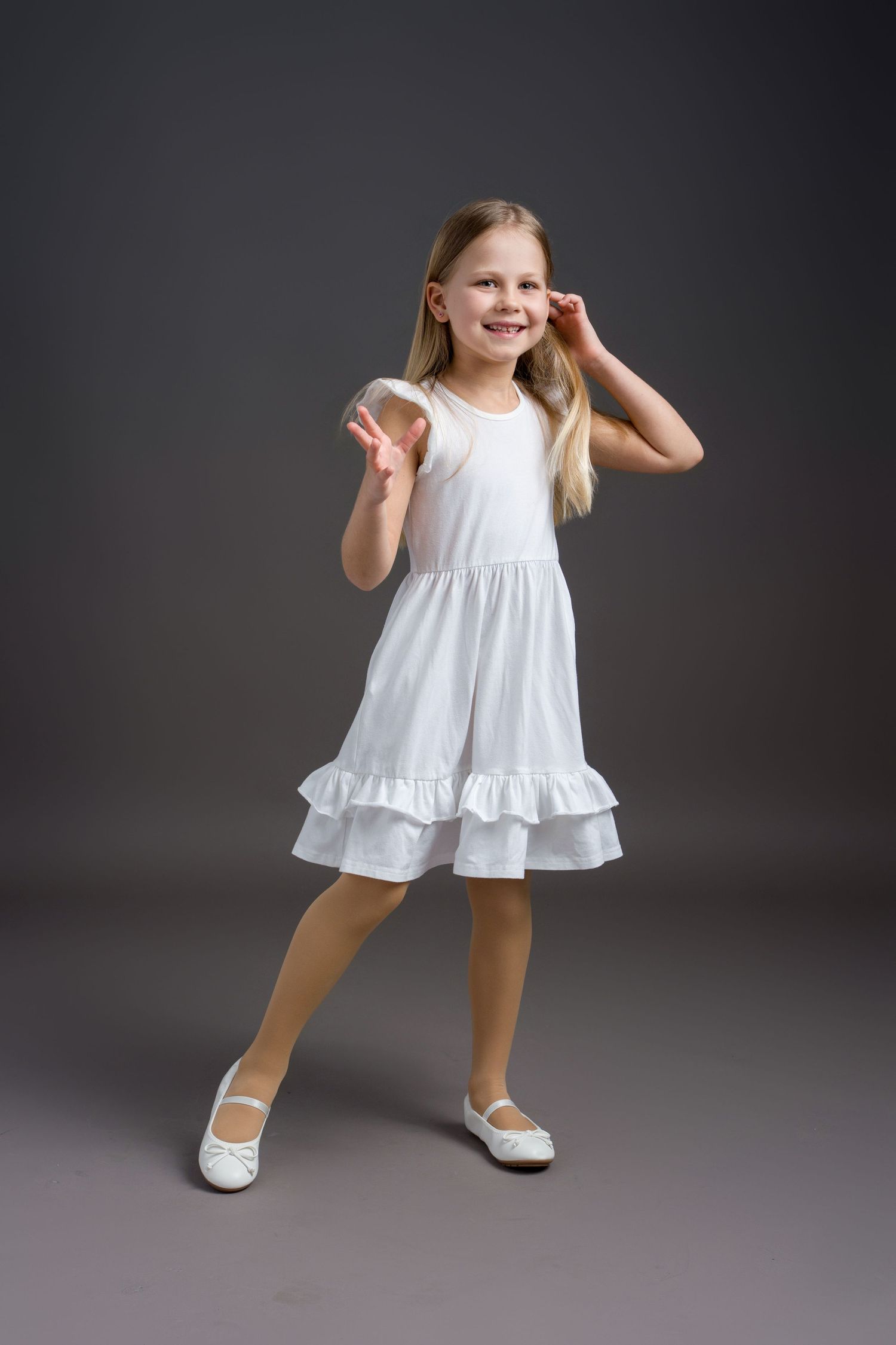 A child in a white ruffled dress poses playfully against a dark studio backdrop.