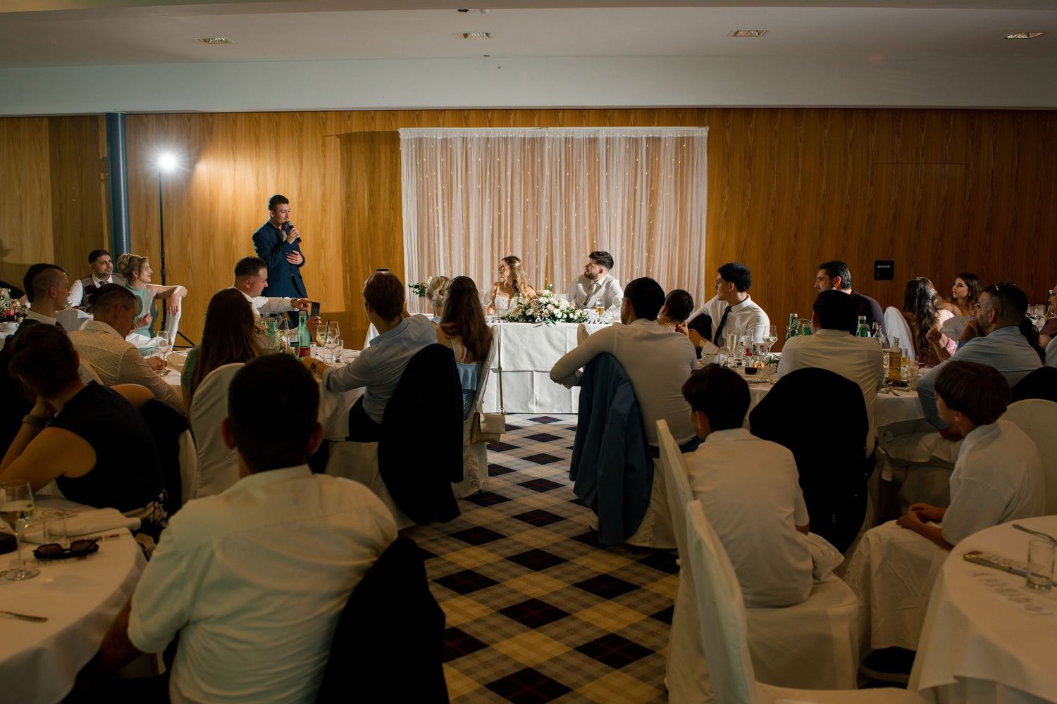 Elegant wedding reception in a banquet hall with guests seated at round tables while a man gives a toast.