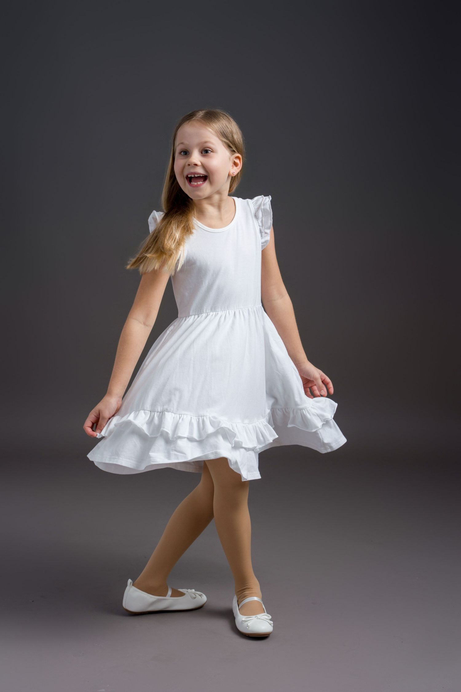 Young dancer in white ruffled dress twirls joyfully against dark backdrop wearing white ballet flats.