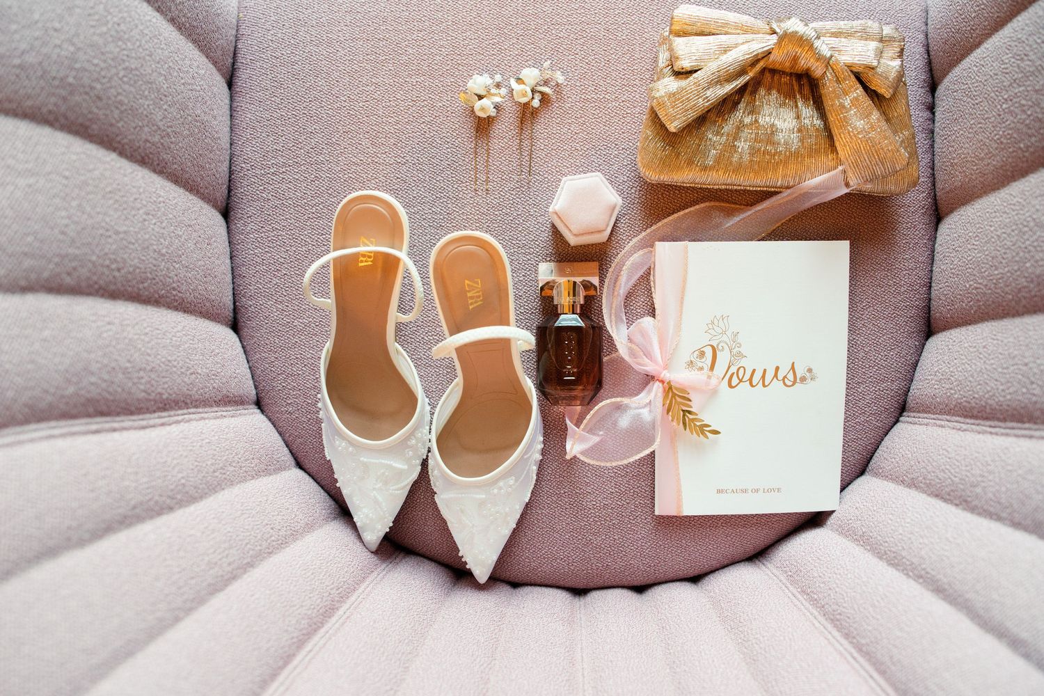 Wedding accessories including white shoes, rings, and invitations arranged on pink fabric.