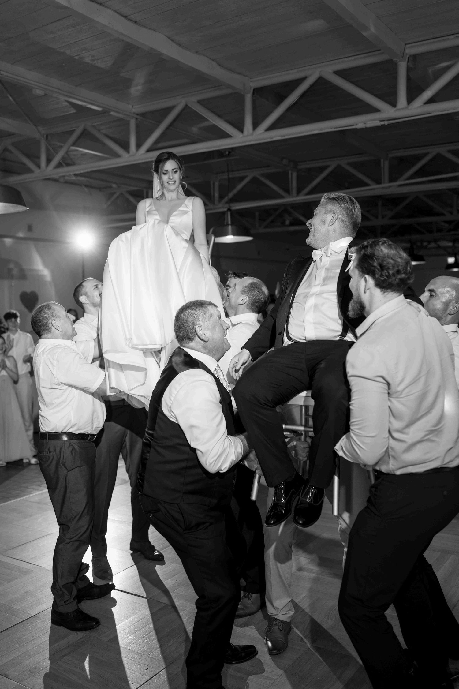 Black and white photo of wedding guests lifting bride and groom during reception dance floor celebration.