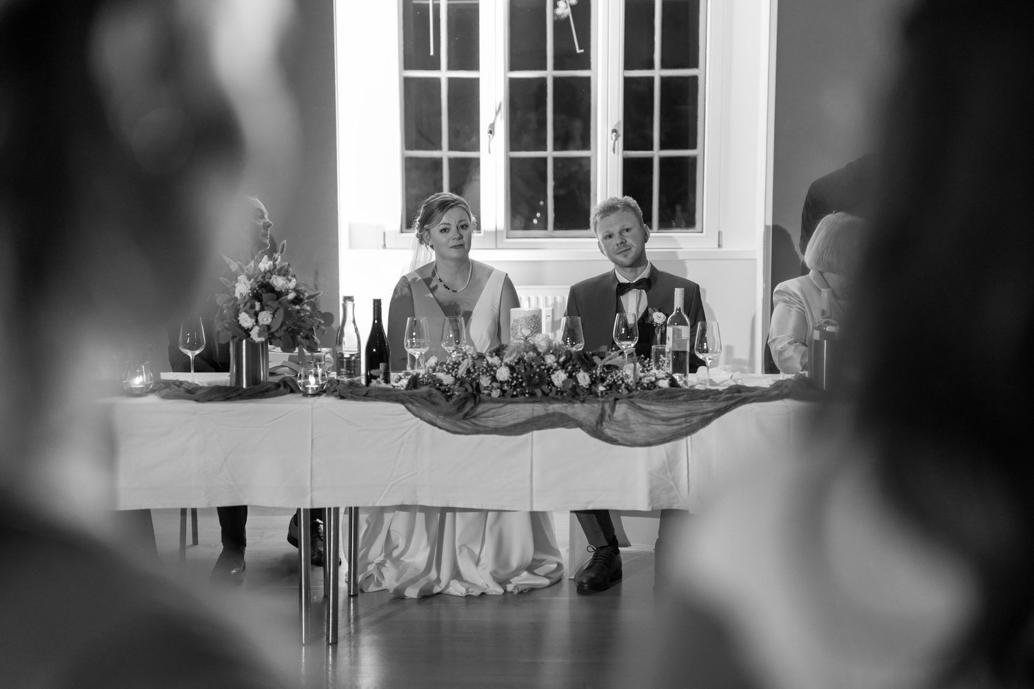 A black and white wedding reception photo showing guests seated at tables in an elegant indoor venue.