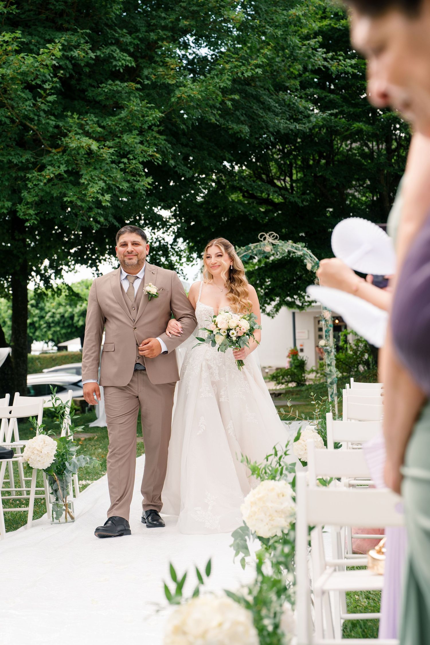 Bride and groom walking down outdoor wedding aisle, bride in white gown holding bouquet, groom in tan suit.