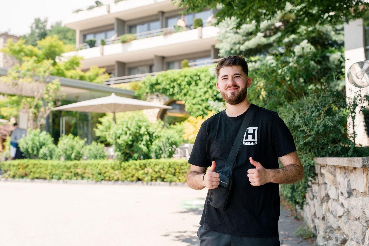 Young smiling man in black t-shirt giving thumbs up outdoors near a modern building with lush green trees.