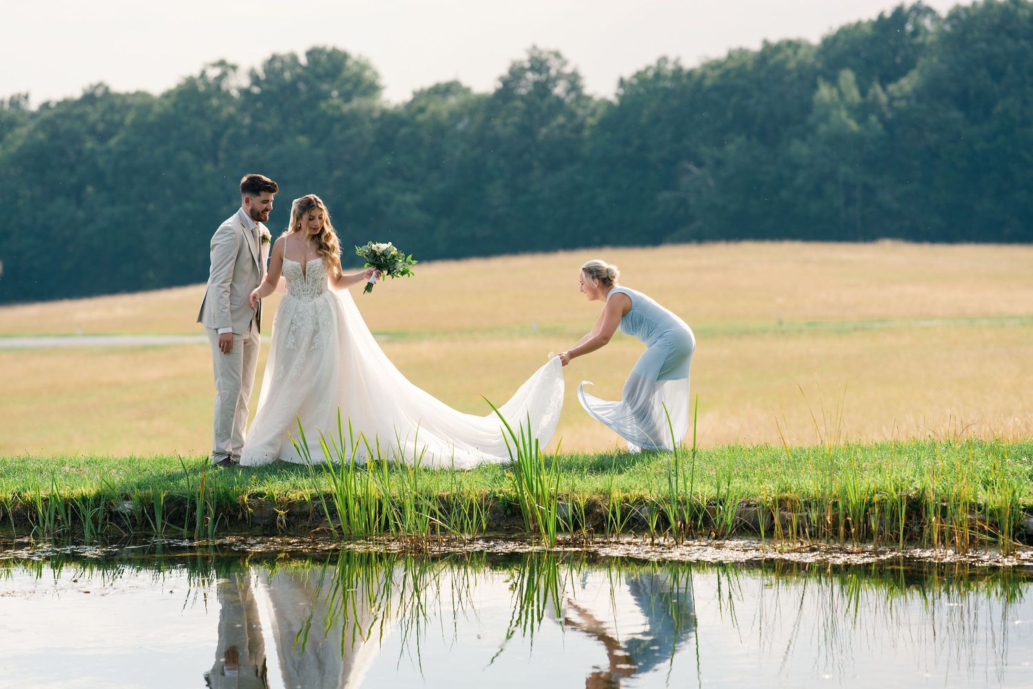Bride and groom stand by a pond while a bridesmaid arranges the wedding dress train in a scenic field.
