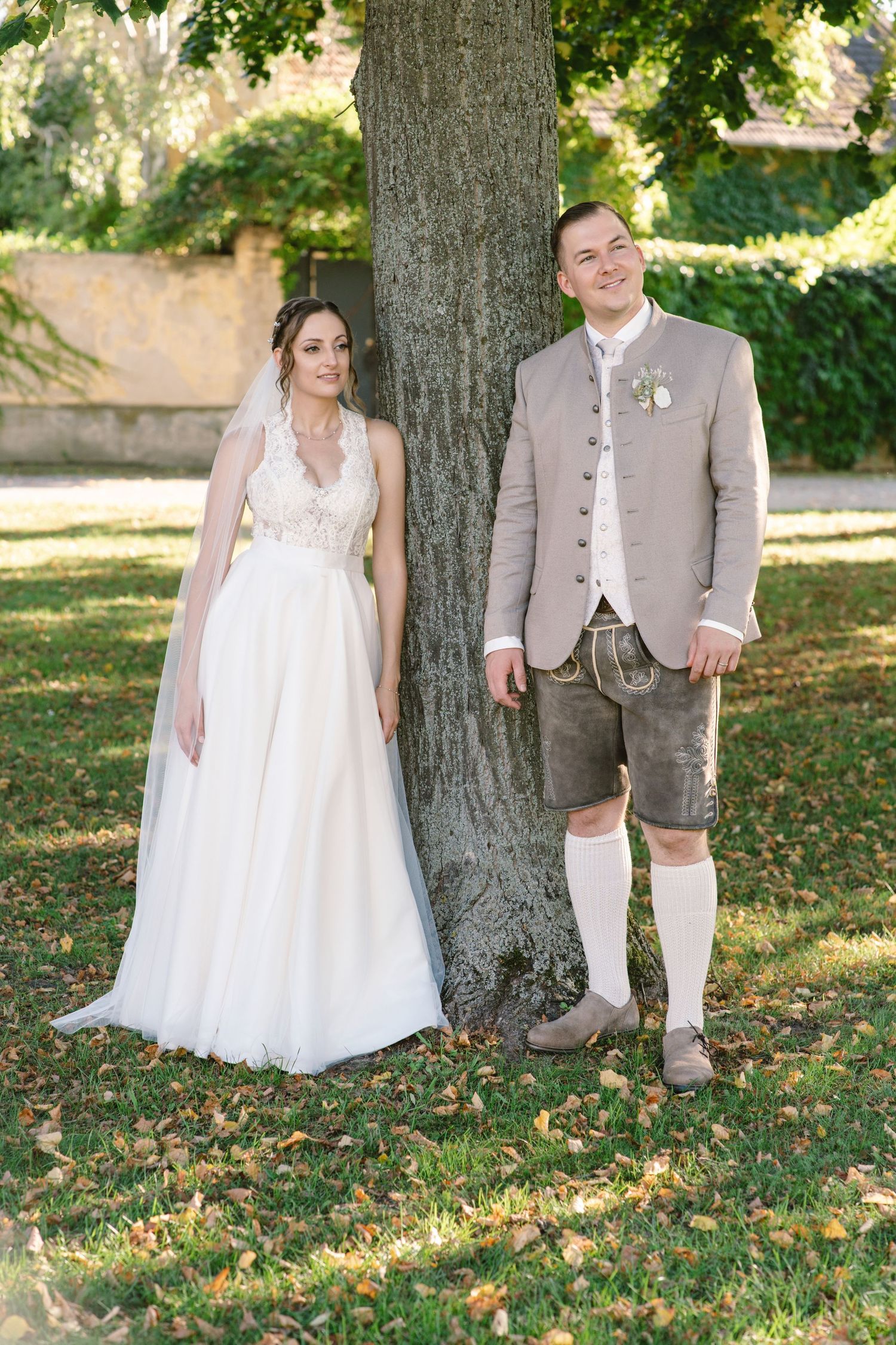 Wedding couple in Bavarian attire poses under a large tree on grassy lawn.