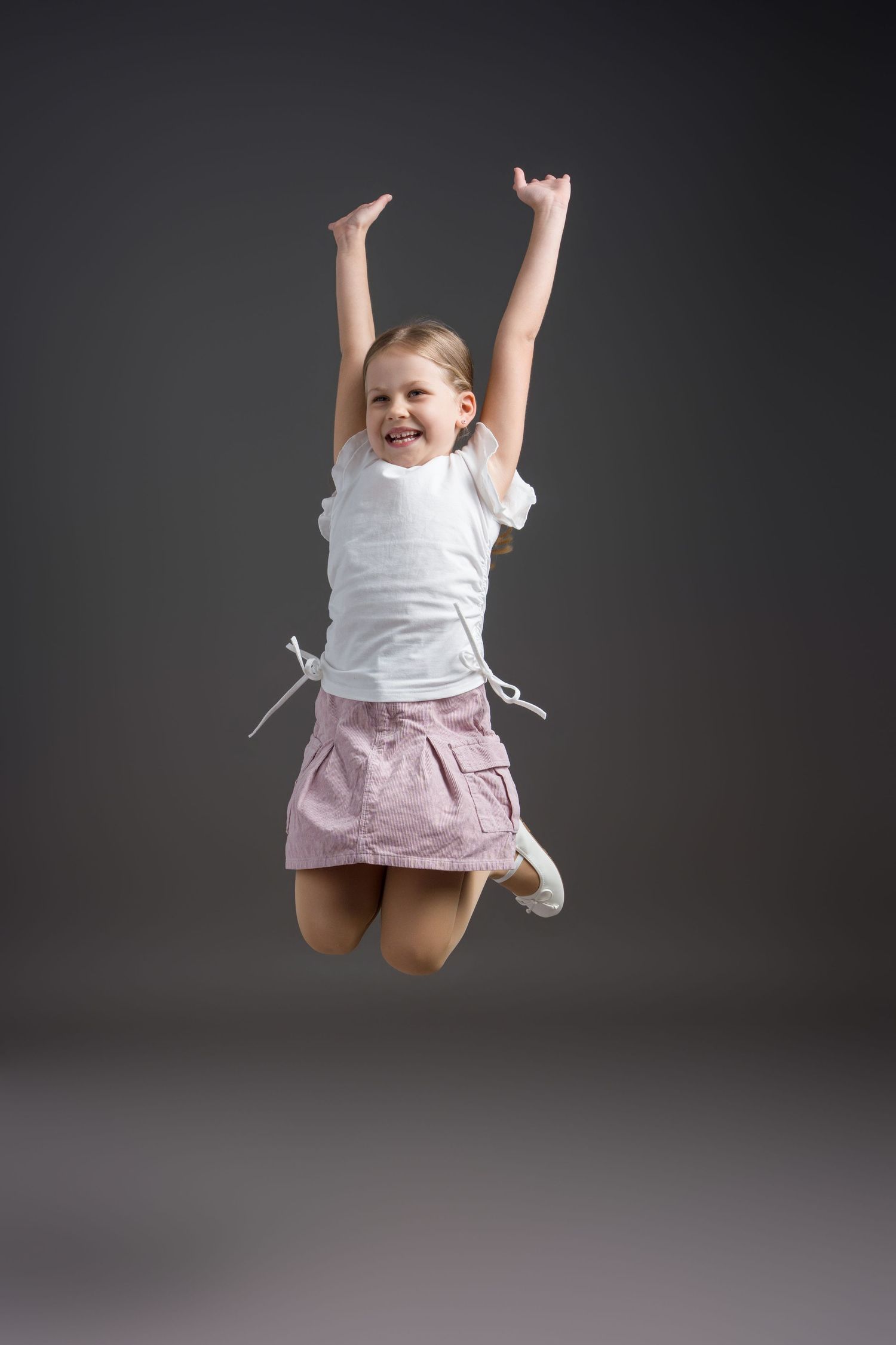 Joyful child in white top and pink skirt jumps high with raised arms against dark backdrop.