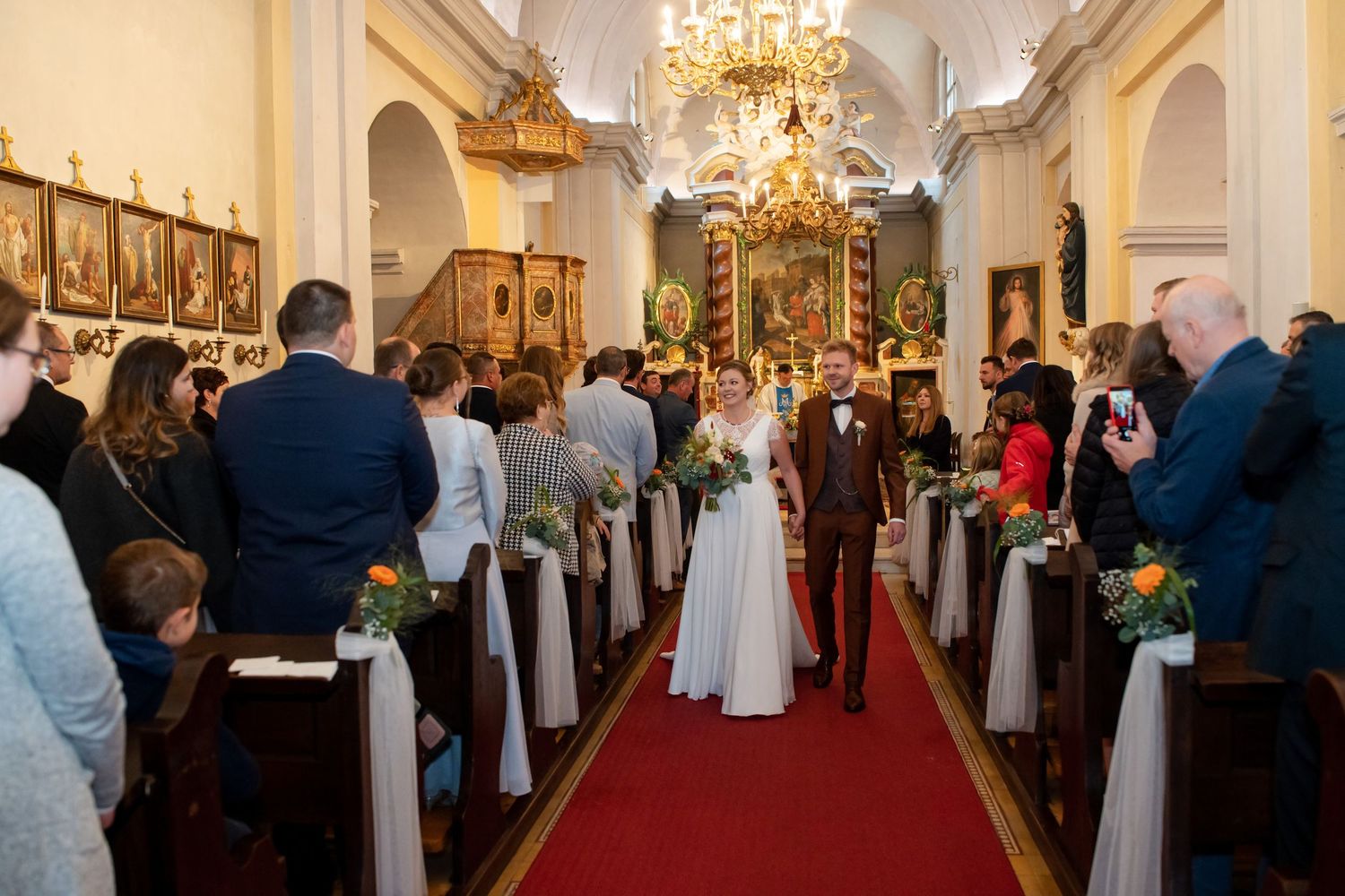 Wedding ceremony in ornate church with chandelier and red carpet aisle.