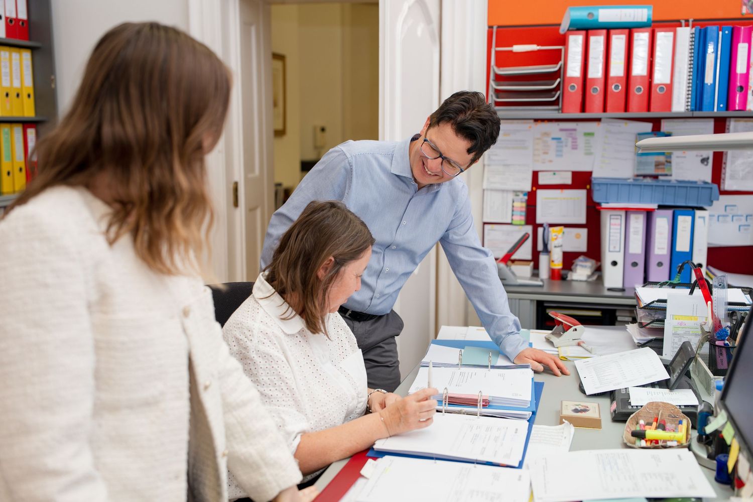 Three office workers collaborating at a desk surrounded by colorful binders and files in a busy workplace.