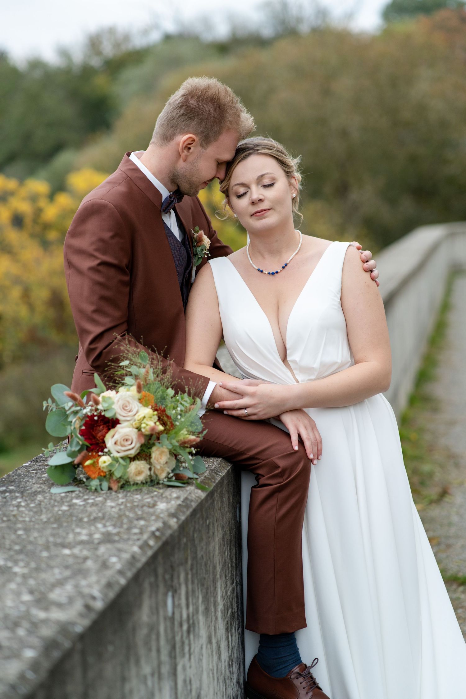 A romantic wedding couple poses on a stone bridge surrounded by autumn foliage and golden flowers.