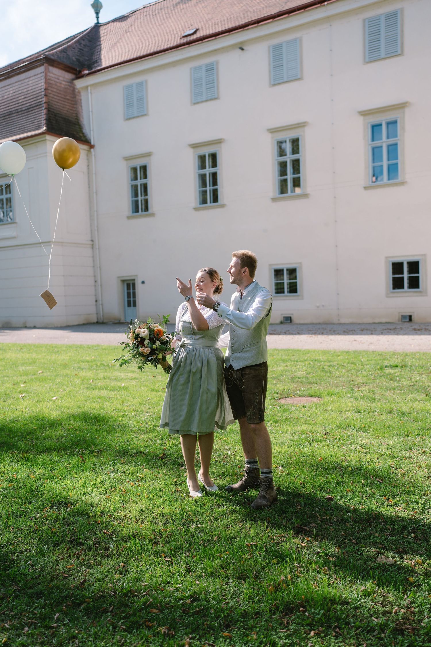 Couple in traditional Bavarian attire playing with balloons on a grassy lawn outside a white building.