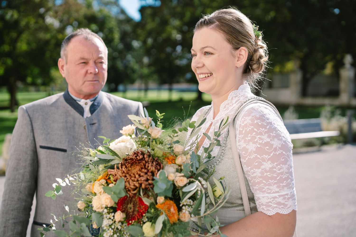 A couple shares a romantic outdoor moment surrounded by greenery while holding autumn-colored floral bouquets.